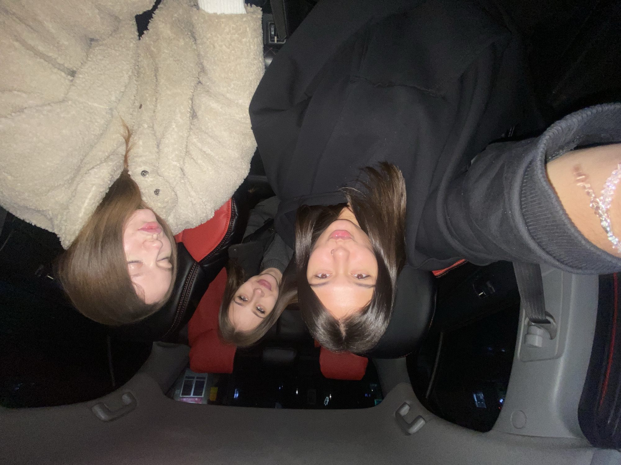 Three young women are in the back seat of a car. The woman on the left is wearing a light-colored, fluffy coat. The woman in the middle is wearing a black jacket. The woman on the right is wearing a black hoodie. The car is driving through the city of Chaikovsky, Russia, at night. There are buildings and streetlights visible outside the window.