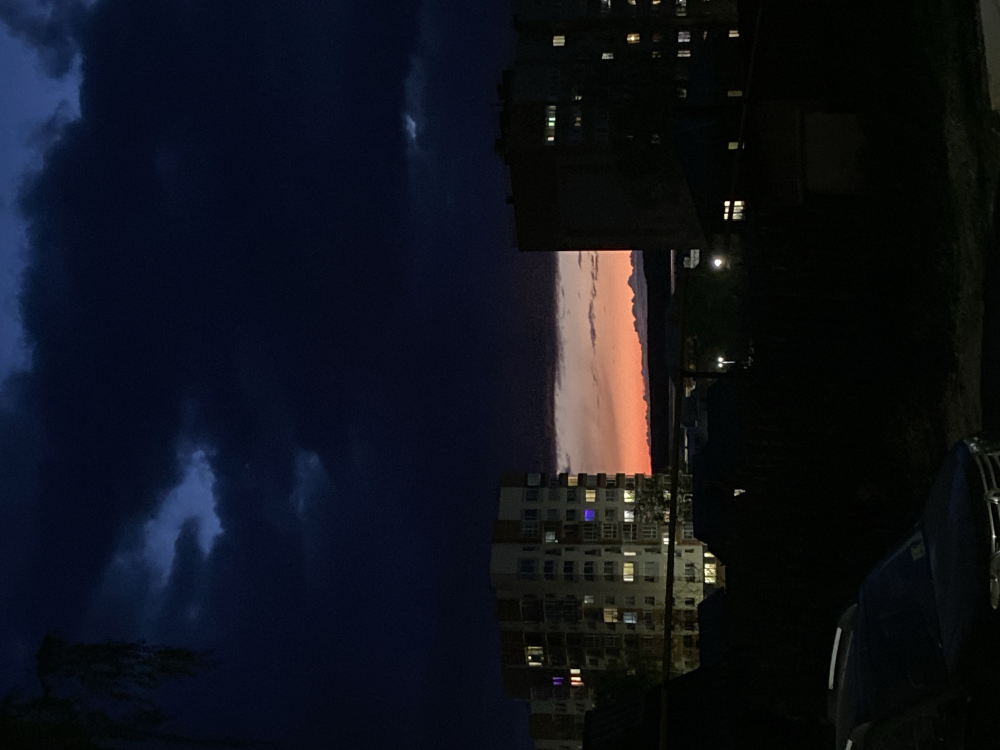 The image depicts a night scene in Votkinsk, Russia, with a dramatic sky divided between dark, stormy clouds and a narrow strip of reddish-orange sunset. Two multi-story apartment buildings flank the sky, their lit windows indicating occupancy. Streetlights are visible, and there are hints of greenery. A dark, indistinct object, possibly a vehicle, occupies the lower right corner of the frame.