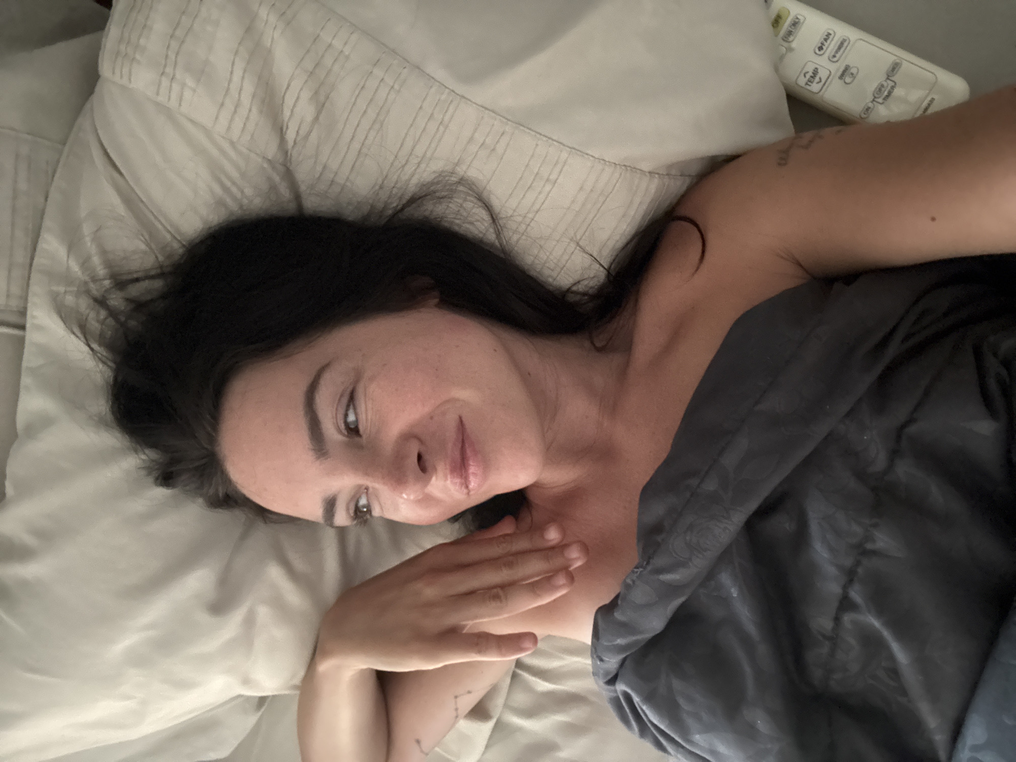 A woman is lying in bed with a grey comforter pulled up to her chest. She has dark hair and is looking to her left with a slight smile. Her left arm is resting on her chest and shoulder, and her right arm is bent and resting on her head. A remote control is visible to her right. The scene appears to be in a bedroom in North Kuta, Indonesia.
