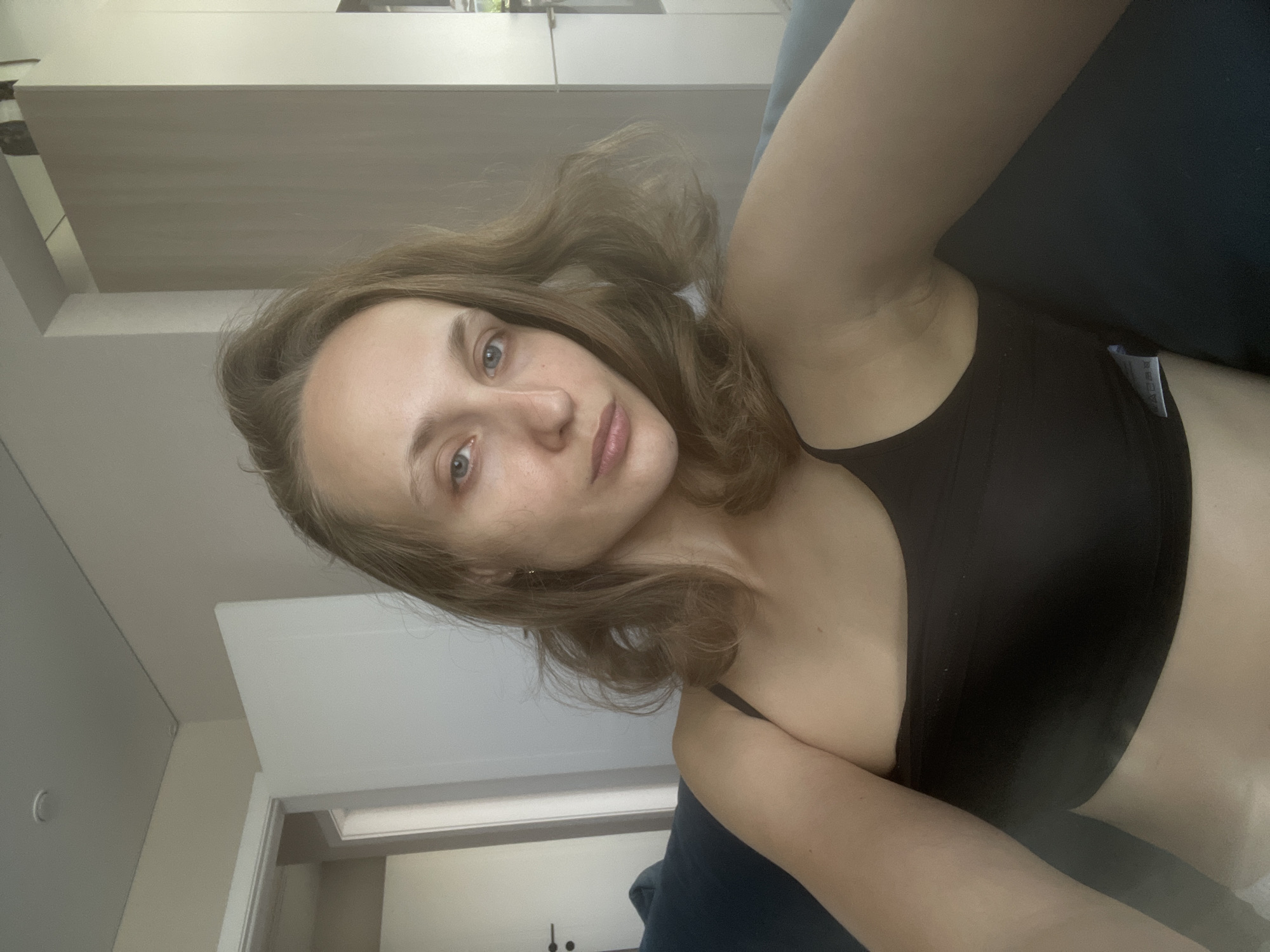 A young woman with fair skin and light brown hair is seated on a dark blue sofa. She is wearing a black one-shoulder tank top. Her gaze is directed towards the camera, and her expression is neutral. The background shows an interior environment with a white door on the left and a large, light-colored cabinet on the right. The setting appears to be a residential interior, possibly an apartment or house. The location specified, Подшивалово, Russia, is a village in the Tver Oblast.