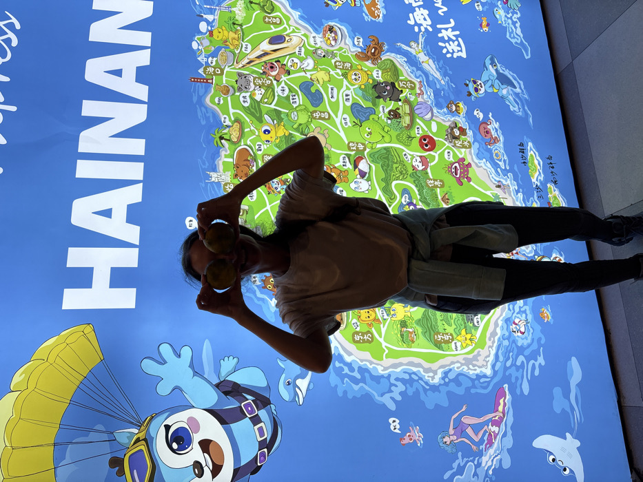 The image displays a single adult female standing indoors in Tianya District, China, positioned directly in front of a large, brightly illuminated wall display. The display features a stylized, vibrant green and blue cartoon map of Hainan Island, with the word "HAINAN" prominently spelled in large white capital letters across the top. The map is adorned with numerous cartoon illustrations, including a blue, deer-like character parachuting in the upper left, a dolphin, and a female character surfing. The female subject, with visible dark hair, wears a light-colored short-sleeved t-shirt, dark fitted pants, and dark athletic shoes; a light-colored long-sleeved garment is tied around her waist. She is holding two round, yellowish-brown objects, possibly fruits, directly in front of her eyes, obscuring them. The subject is smiling with her mouth open. The floor beneath her consists of light-toned tiles.