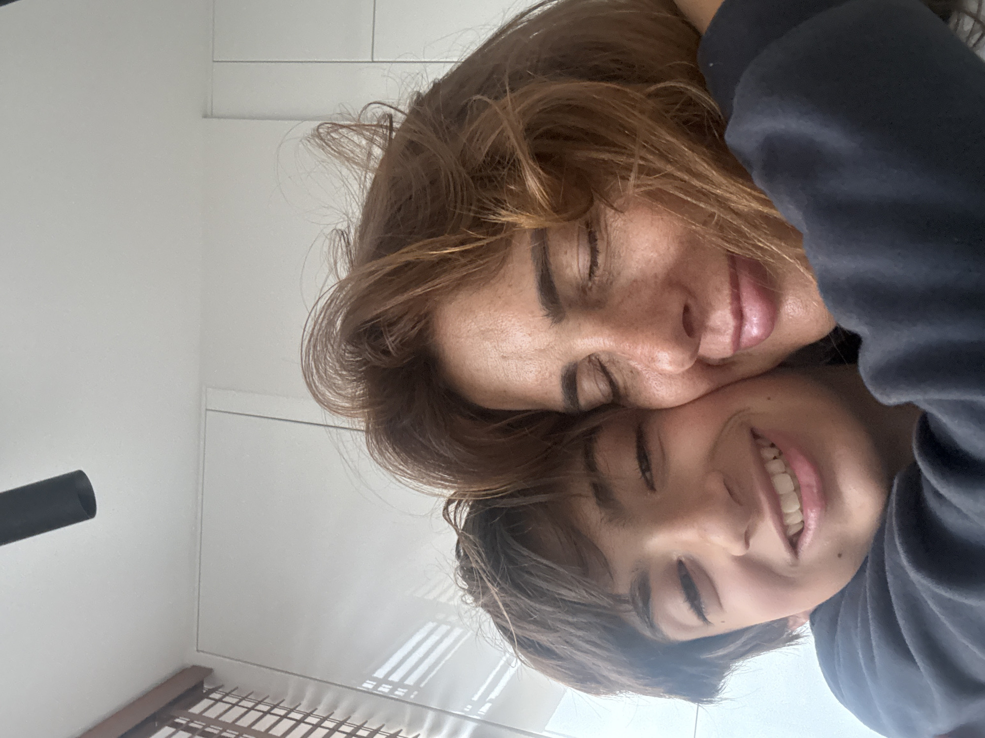 The image shows two people, presumably a mother and child, close together with their faces near each other. The woman has brown hair and freckles, and her eyes are closed. The child is smiling, and their eyes are also closed. They are wearing a dark-colored shirt or sweater. In the background, there is a white wall, and the corner of a bed is visible, as well as what appears to be a black tube. Based solely on the provided information, it is impossible to determine if the image was taken in Vila Nova de Gaia, Portugal, or any other location.