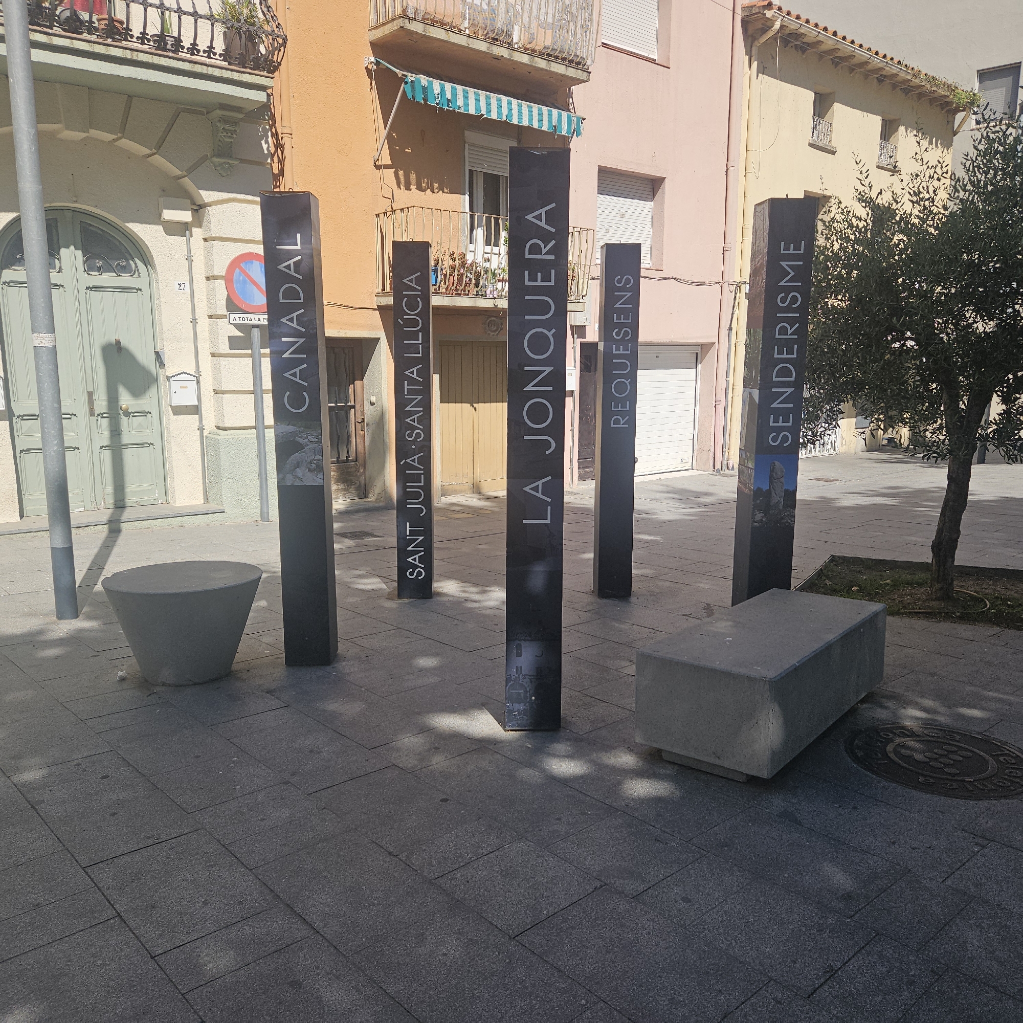 The image shows an outdoor public square in La Jonquera, Spain. Five vertical, dark-colored signs are arranged in a semi-circle. Each sign displays a name in white lettering: "CANADAL", "SANT JULIA·SANTA LLÚCIA", "LA JONQUERA", "REQUESENS", and "SENDERISME". There are two concrete benches in the square, one near the "CANADAL" sign and another near the "SENDERISME" sign. A small tree is planted near the "SENDERISME" sign, and a manhole cover is visible on the ground nearby. Buildings with varied colors (orange, pink, cream) are in the background. The ground is paved with gray rectangular blocks. A street lamp is visible on the left side of the image. The scene appears to be well-lit, suggesting a sunny day. There is no person shown in this image.