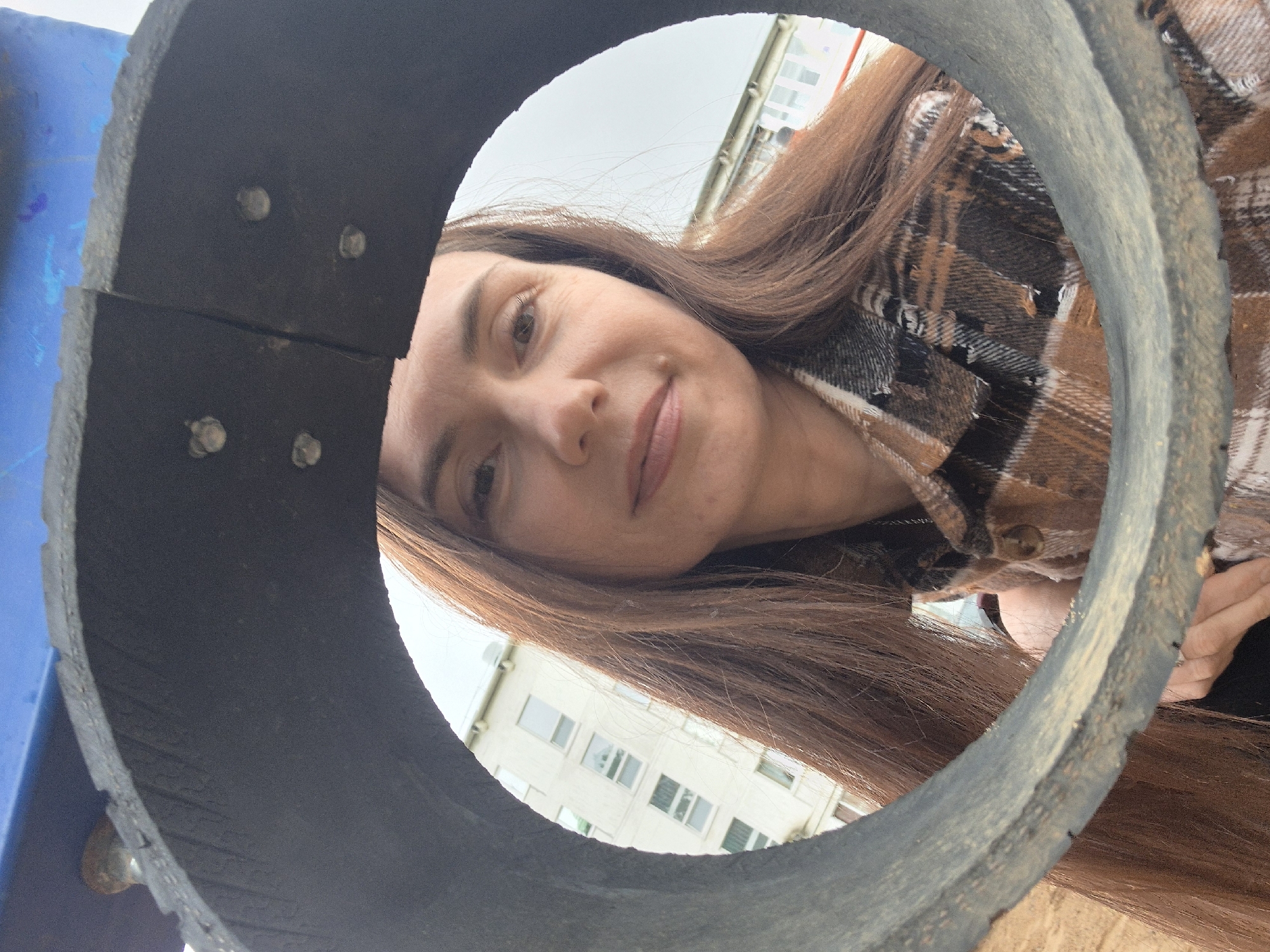 A woman with long brown hair, wearing a plaid shirt, is peering through a dark, circular object, possibly a section of a pipe or tunnel. The object is weathered and appears to be made of a composite material held together by metallic rivets. Part of a white building with multiple windows can be seen in the background, indicating an urban environment. The camera angle is low, suggesting the image was taken from beneath the circular object.