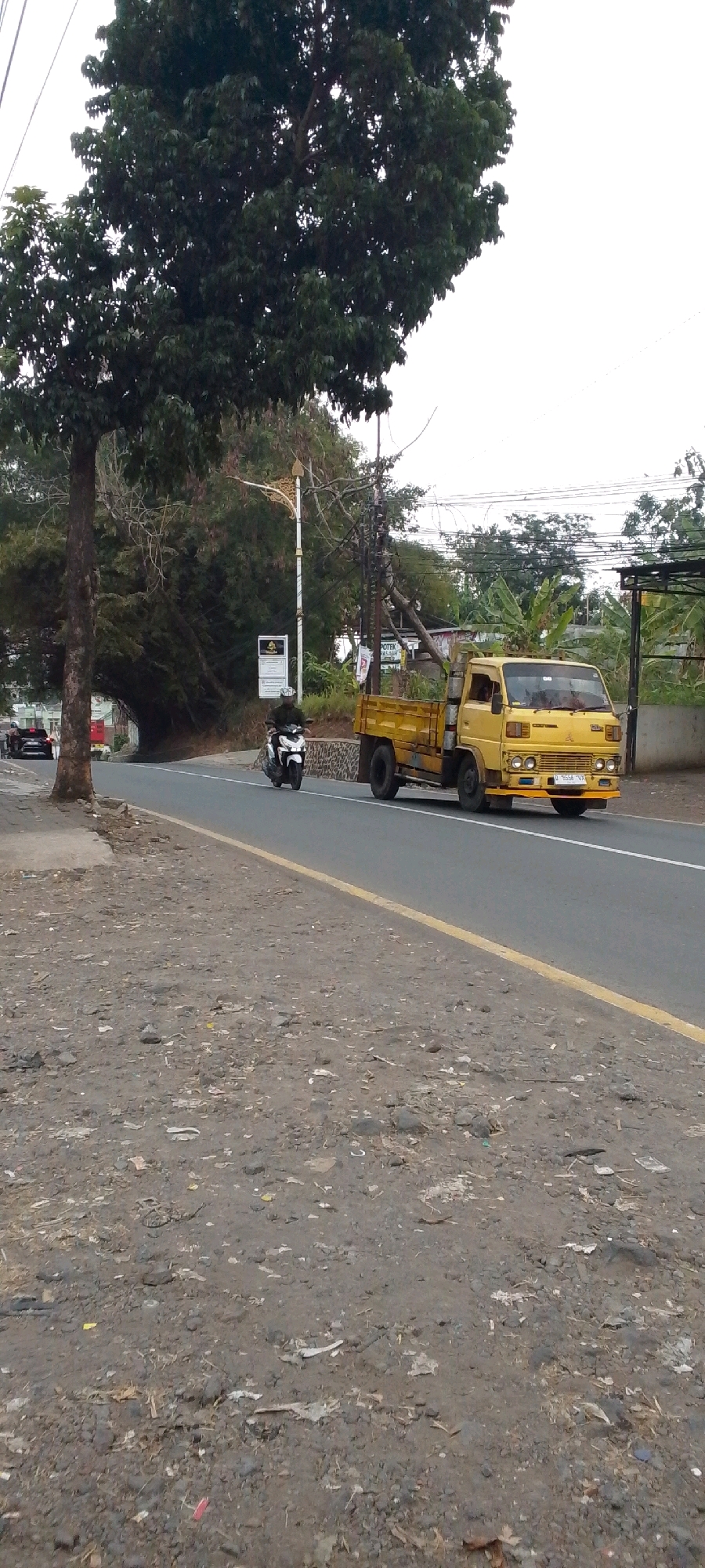 In the city of Cilaku, Indonesia, a yellow flatbed truck, possibly a Mitsubishi Colt, is traveling on an asphalt road. The truck's bed is loaded with several light-colored rectangular objects, likely building materials or other cargo. A white motorcycle with a rider is approaching the truck from the left, also on the road. The road has a white lane divider and a yellow line on the right edge. The environment consists of a dirt shoulder with scattered debris, a large tree with dark green foliage on the left, and a line of trees and vegetation in the background. A lamppost with ornate gold accents stands on the left side of the road, near a sign. Another sign is visible further down the road. The sky is overcast.