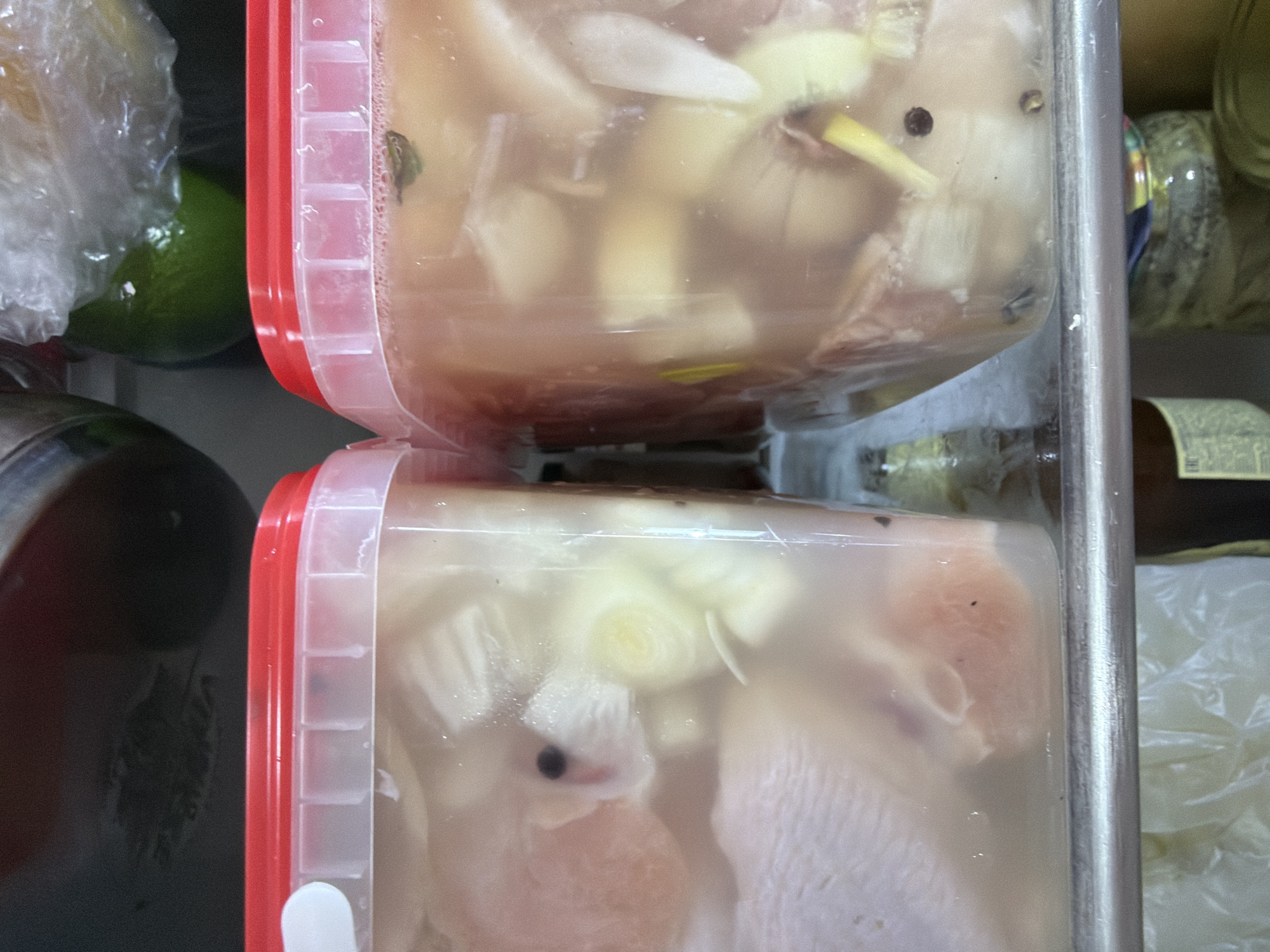 Two clear plastic food containers with vibrant red lids are stacked vertically, dominating the center of the image. Both containers are filled with a translucent, milky liquid, suggesting brine or marinade, and contain raw food items.
In the upper container, large pieces of light-colored raw meat, possibly poultry or pork, are visible, submerged in the liquid. Alongside the meat are chunks of white onion, slices of a yellowish-green vegetable that resembles ginger or leek, and scattered black peppercorns. Bubbles cling to the interior sides of the container, indicating the presence of air or a reaction.
The lower container mirrors the contents of the upper one, showing similar cuts of raw meat, white onion, and peppercorns. A distinct round slice of white onion, possibly a cross-section of a scallion bulb, is visible near the front. The meat appears pinkish-white and uncooked.
To the left of the upper container, a bright green lime, partially wrapped in clear plastic, is visible, resting on a dark surface. Below it, a metallic object, possibly part of a pot or container, is partially seen. To the right of the containers, in the background, various bottles and jars are stored on what appears to be a shelf, their labels and contents mostly indistinct due to blur and poor lighting, though one bottle has a dark brown liquid. The overall setting appears to be an indoor kitchen or food storage area, characterized by utilitarian containers and food items. No discernible text is legible on any of the containers or background items. The image is taken from a slightly upward angle, looking down on the stacked containers. The lighting is diffused and appears to be artificial, common for an indoor setting.