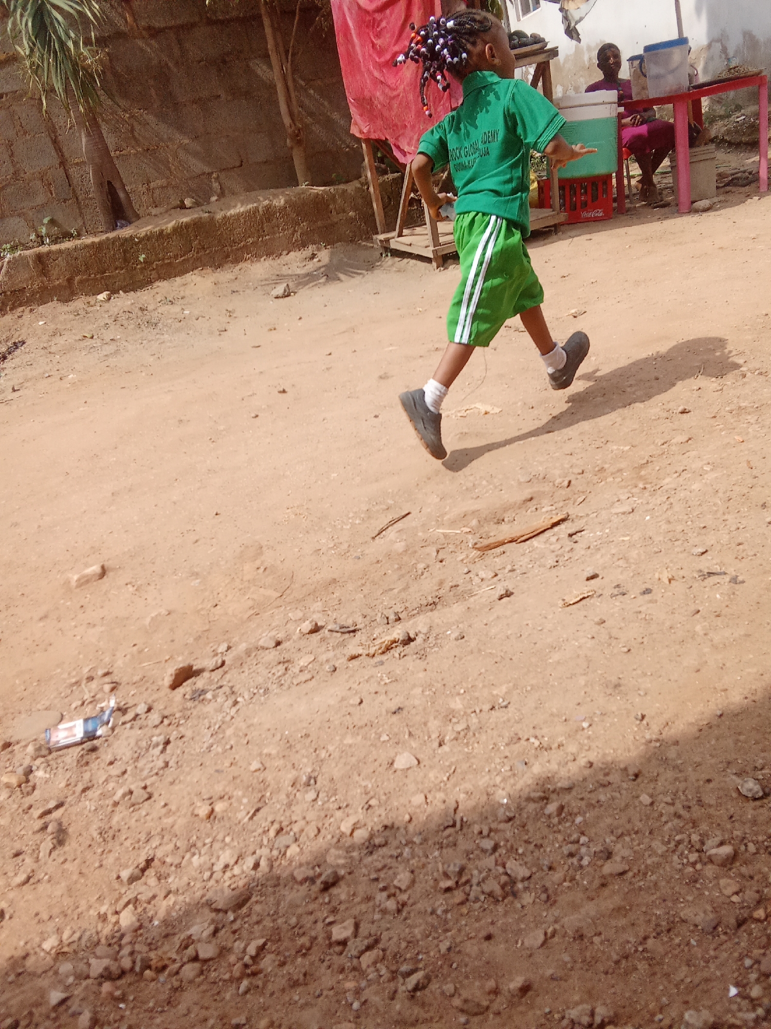 A young child with braided hair adorned with colorful beads runs across a dirt path in Kagini, Nigeria. The child is wearing a green polo shirt with the inscription "ROCK GLOBAL ACADEMY ABUJA" and green shorts with white stripes. The child is also wearing white socks and dark shoes. In the background, a person sits on a chair behind a table. A red tarp is visible behind the child, and a wall made of cement blocks forms the backdrop. The ground is covered in dirt and small stones. The scene appears to be outdoors, possibly in a rural or semi-urban area.