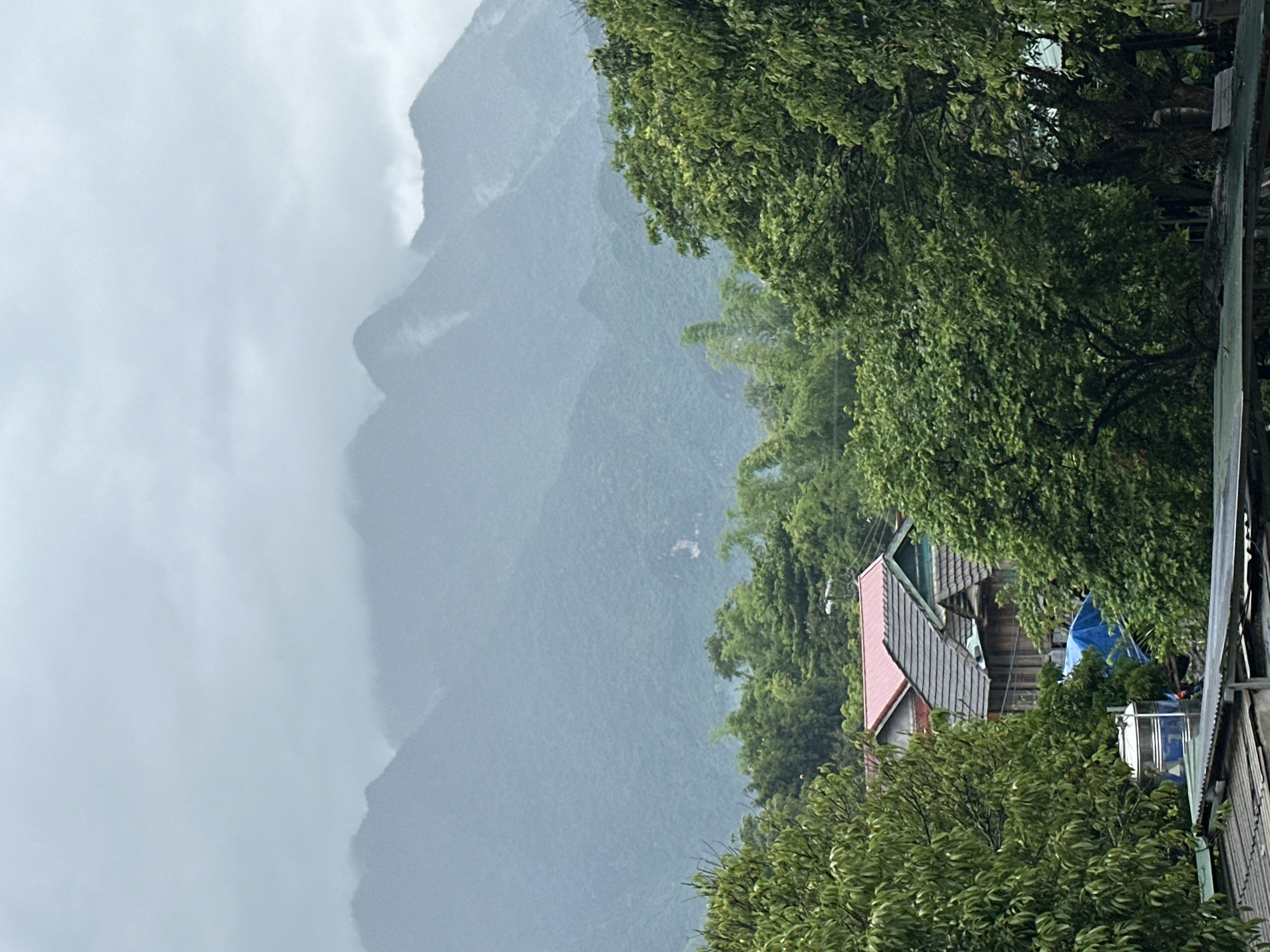 This is a photograph taken from a slightly elevated vantage point, looking out over a village nestled at the foot of a mountain range. The sky is overcast, with dense grey clouds obscuring the tops of the mountains, which are covered in lush green vegetation. Several distinct peaks can be seen, their slopes appearing steep and rugged.
In the mid-ground, the village of Xóm Khán, Vietnam, is visible. Traditional houses with tiled roofs, some red and others dark grey, are interspersed with dense foliage. One house appears to have wooden siding and a dark, multi-tiered roof. A bright blue tarp is spread out near some of the houses, and a silver water tank with a blue base is also visible. The trees in the foreground are tall and leafy, framing the scene on both sides. Bamboo stalks are also present amongst the trees. The overall impression is one of a rural, somewhat misty landscape, likely during or just after a period of rain. There are no people or discernible actions visible in the photograph.