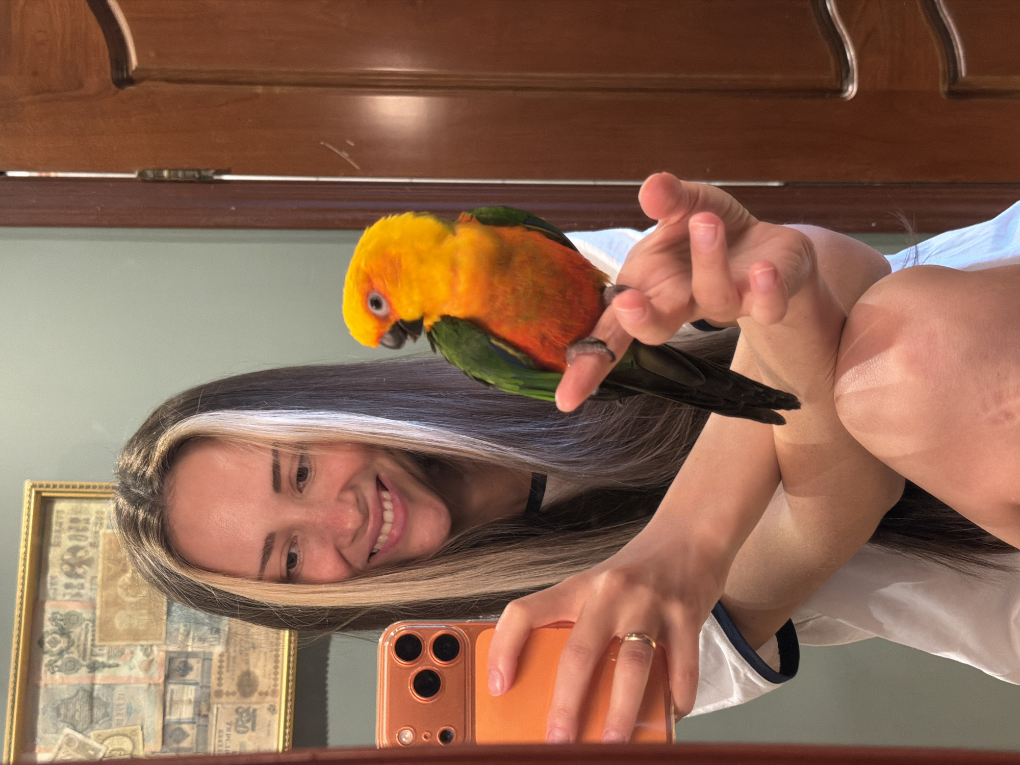 A young woman with dark hair with blonde streaks is taking a selfie in a mirror. She is smiling and holding an orange smartphone in her left hand. A colorful parrot, with predominantly yellow and green plumage, is perched on her right index finger. The woman is wearing a white t-shirt with navy blue trim. She has a ring on her left ring finger. In the background, a framed artwork hangs on a pale green wall, and a wooden door is visible to the right. The scene appears to be indoors, possibly in a home. There is no visible indication that the location is in Khimki, Russia.