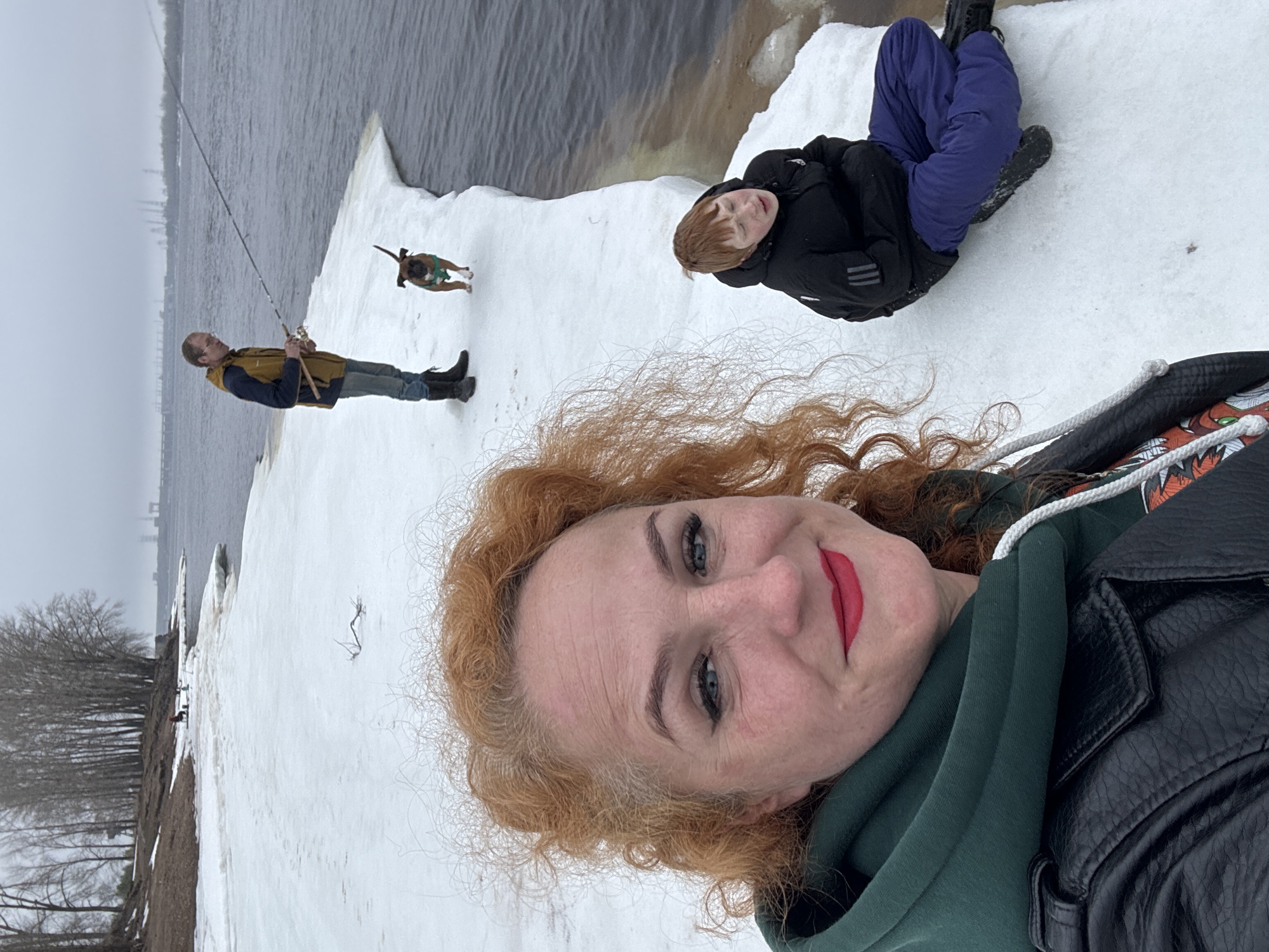 The image shows a woman taking a selfie outdoors. She has red hair, blue eyes, and is wearing red lipstick, a black leather jacket, and a green hooded sweatshirt. Behind her, a man is fishing next to the river. Next to him is a dog. A child is lying on the snow. The setting appears to be snowy and overcast, near a body of water. The picture was taken in the city of Chaikovsky, Russia.