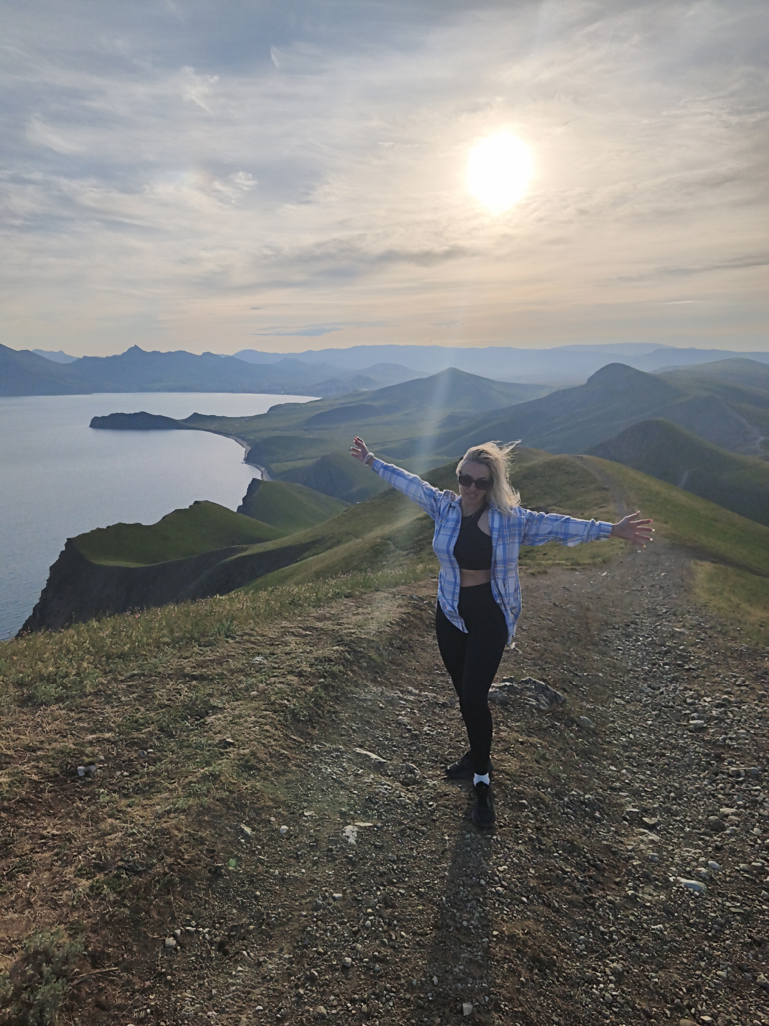 A woman stands atop a rocky hillside overlooking the sea and distant mountains. She wears a black sports bra, black leggings, a blue and white plaid shirt, and sunglasses, with her arms outstretched. The sky is bright with sunlight, filtered by thin clouds. The hillside consists of low vegetation and rocks. The sea stretches out to the left, bordered by green hills and cliffs. The background features layered mountains fading into the horizon. The location is the city of Ordzhonikidze, Ukraine.