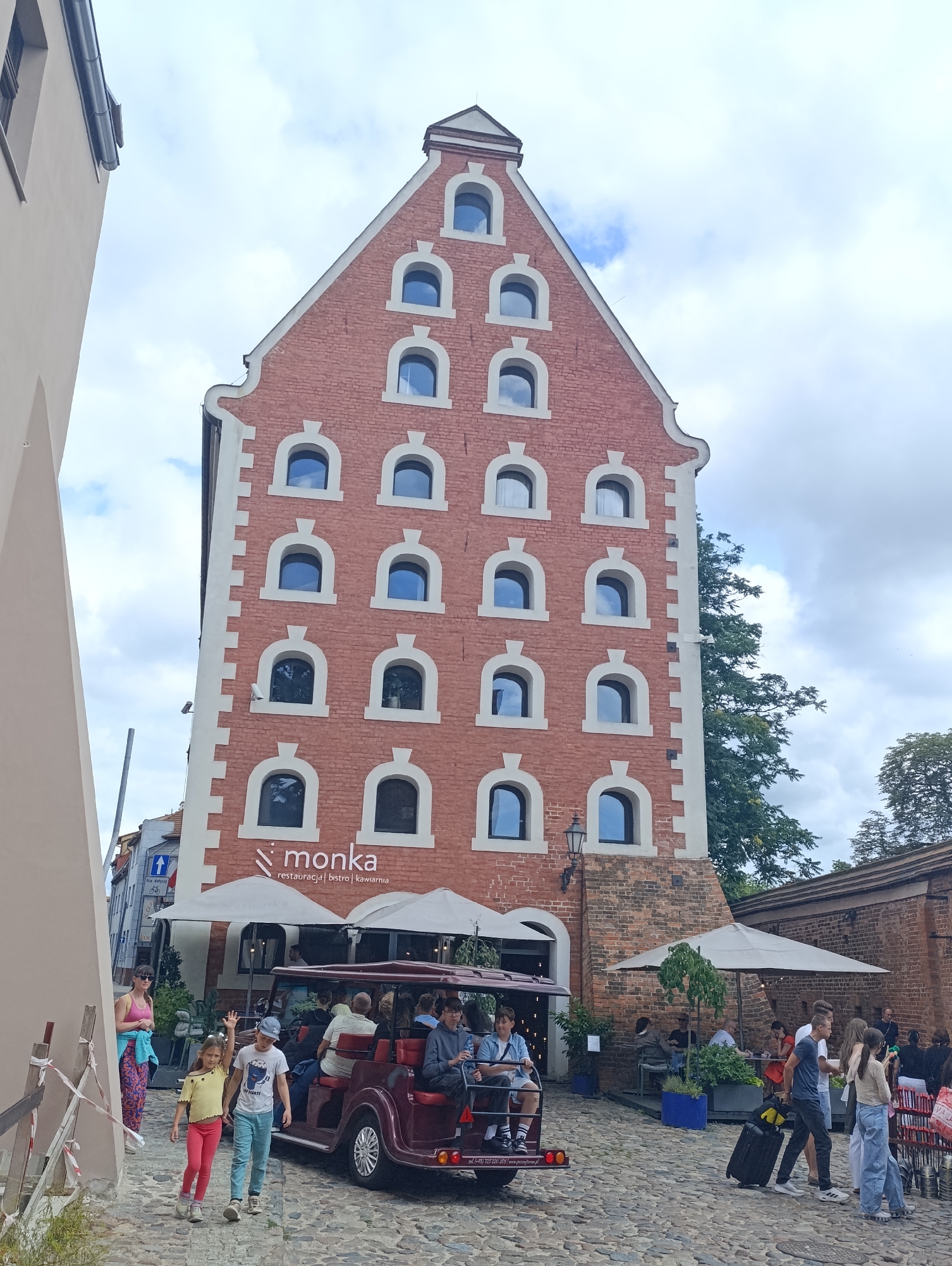 The image captures a scene on a cobblestone street in Toruń, Poland, likely near the historic Old Town. Dominating the frame is a tall, narrow brick building with a distinct stepped gable roof, characteristic of Hanseatic architecture. The building facade is adorned with numerous arched windows with white surrounds, arranged in a grid pattern. Below, the building houses a restaurant or cafe identified by the "Monka restauracja | bistro | kawiarnia" sign. Outdoor seating with umbrellas is set up in front of the establishment.
In the foreground, a group of people are present. Two children, a girl in a yellow top and red pants, and a boy in a grey t-shirt and blue pants, are walking towards the viewer. The girl is waving. Behind them, a red, open-air tourist vehicle, resembling a small train or trolley, carries several passengers. Further back, other individuals are seen walking, some with luggage, and seated at the outdoor tables. The background features more brick buildings and trees, under a cloudy sky. The overall impression is a bustling urban environment, possibly a tourist area.