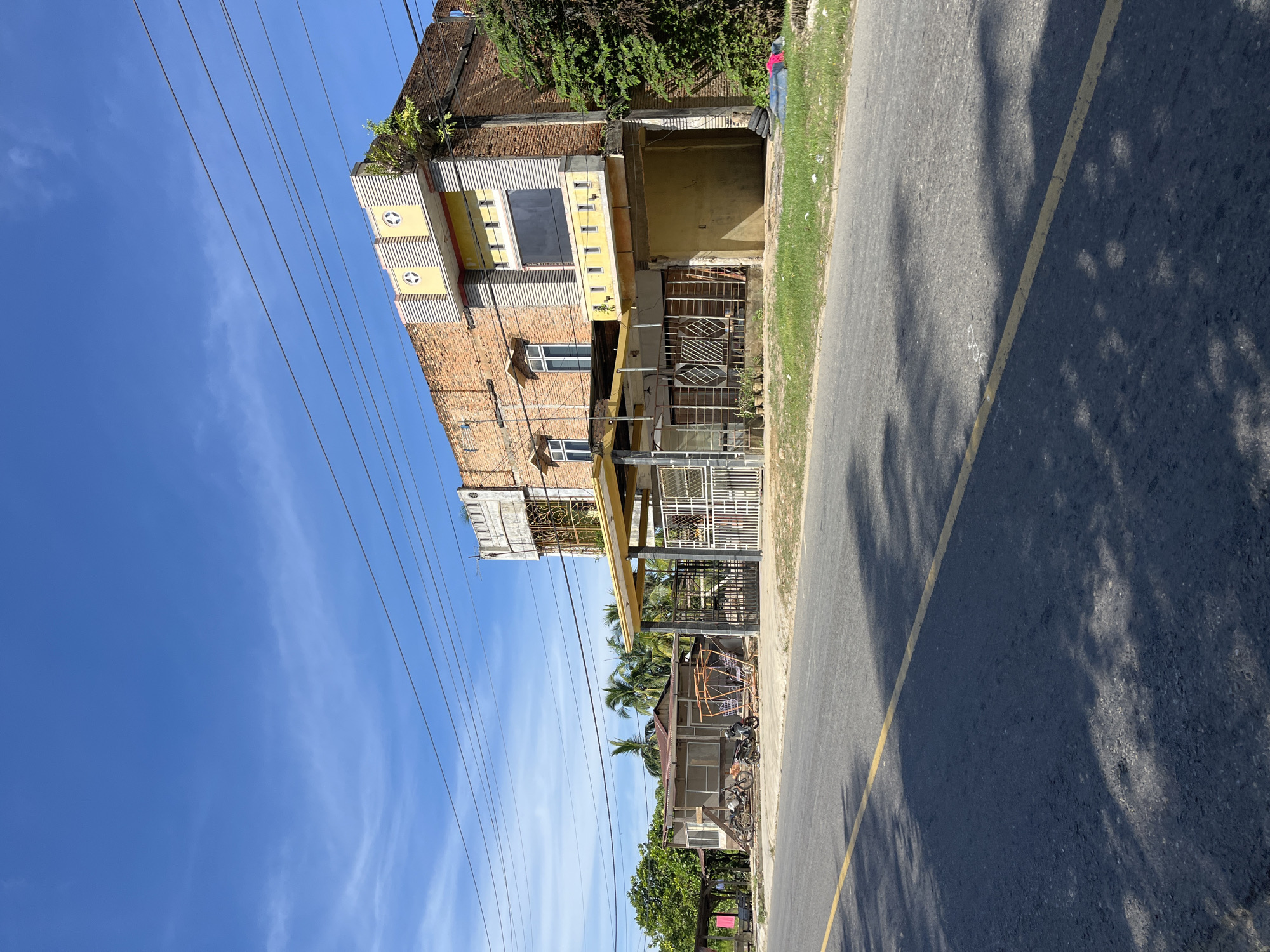 A street scene in Pangkalan, Indonesia, features a two-story brick building with some construction elements, a covered porch, and a fenced entrance. In front of the building, a paved road runs horizontally across the foreground with a yellow line marking the center. Trees and smaller structures are visible to the left of the main building. Power lines crisscross the sky above. The sky is bright blue with scattered white clouds.