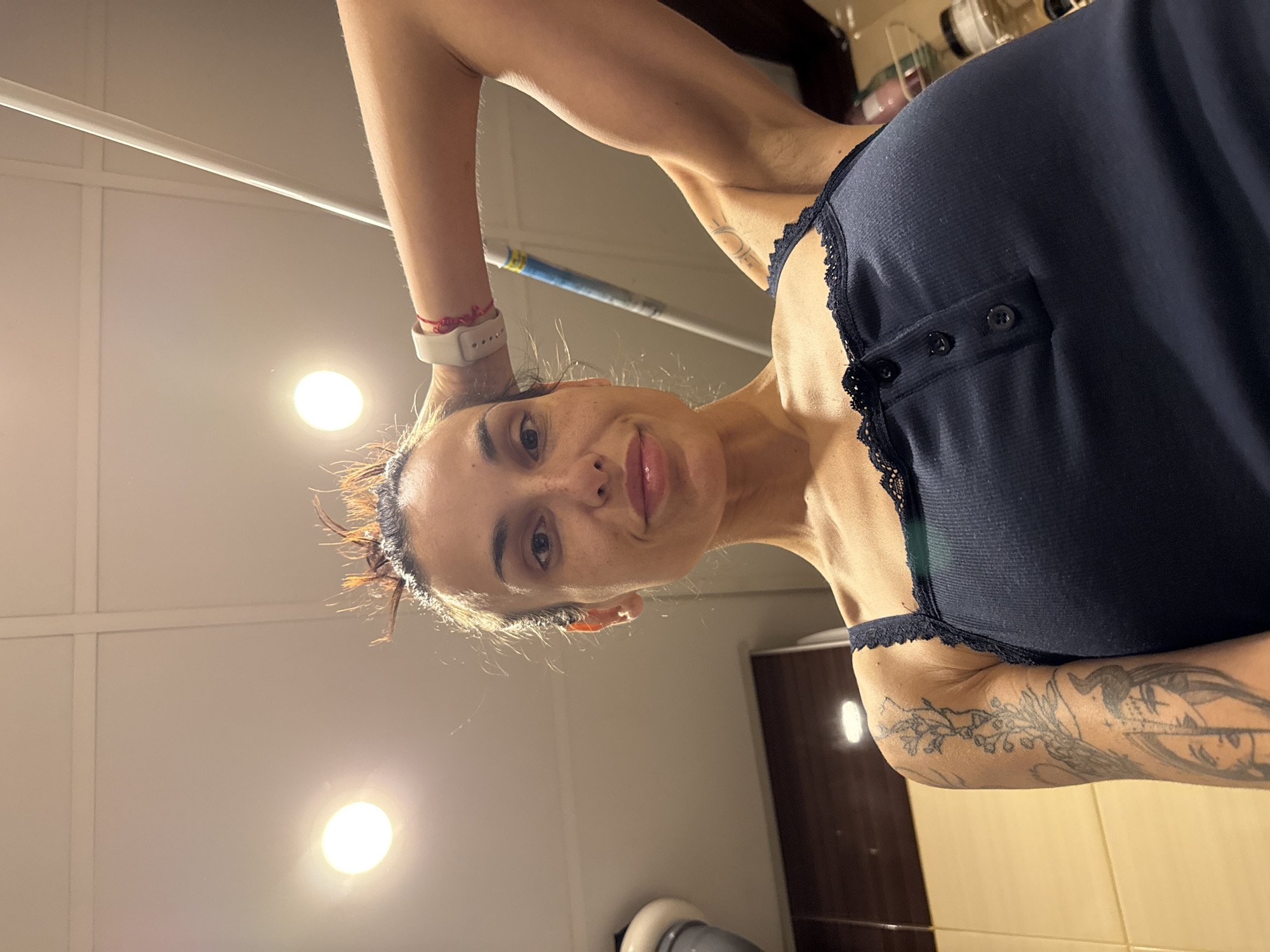 The image is a low-angle selfie of a single adult female in an indoor setting. The individual has dark hair with some reddish highlights, styled upwards with loose strands. She has fair skin with visible freckles on her face and is looking directly at the camera with a slight smile. She wears a dark blue, ribbed tank top with black lace trim along the neckline and straps, featuring three black buttons on the front placket. A white smartwatch and a red braided string bracelet are visible on her left wrist, which is bent with the hand placed behind her head. Tattoos are present on both arms; a floral design and a split-face motif (one half appearing natural, the other resembling a horned figure) are visible on her left arm, while a small crescent moon-like tattoo is on her upper right arm.
The background consists of a light-colored ceiling with visible grid lines, featuring two bright, circular overhead lights. To the left, dark brown wall cabinets and light-colored wall panels are partially visible, along with the top of a light-colored object. A white rod extends diagonally from the upper right side of the frame, and blurry shelves holding various containers are visible on the far right. The environment suggests a domestic space, possibly a kitchen or an area with kitchen elements. The image contains no discernible visual information or metadata to confirm a specific geographical location such as the city of Pleven, Bulgaria.