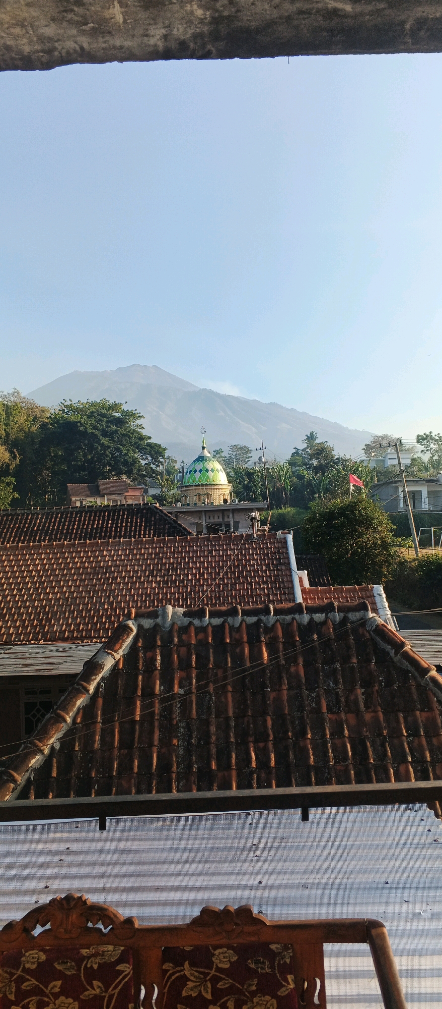 This is a photograph taken from an elevated perspective, looking out over a village landscape. In the foreground, the tiled roofs of several houses are visible, some appearing older and weathered with moss. Beyond the rooftops, a mosque with a distinctive green and blue patterned dome stands prominently. In the background, a large, misty mountain dominates the horizon under a clear, pale blue sky. Lush green trees and foliage surround the buildings, suggesting a rural or semi-rural environment. A red flag is visible on a pole to the right of the mosque. The scene appears to be set in Bocek, Indonesia, given the architectural style and natural scenery. The bottom portion of the image shows a piece of carved wooden furniture with a patterned fabric cushion, implying the photo was taken from indoors looking out. The overall lighting suggests it is daytime, possibly morning or late afternoon due to the soft light.