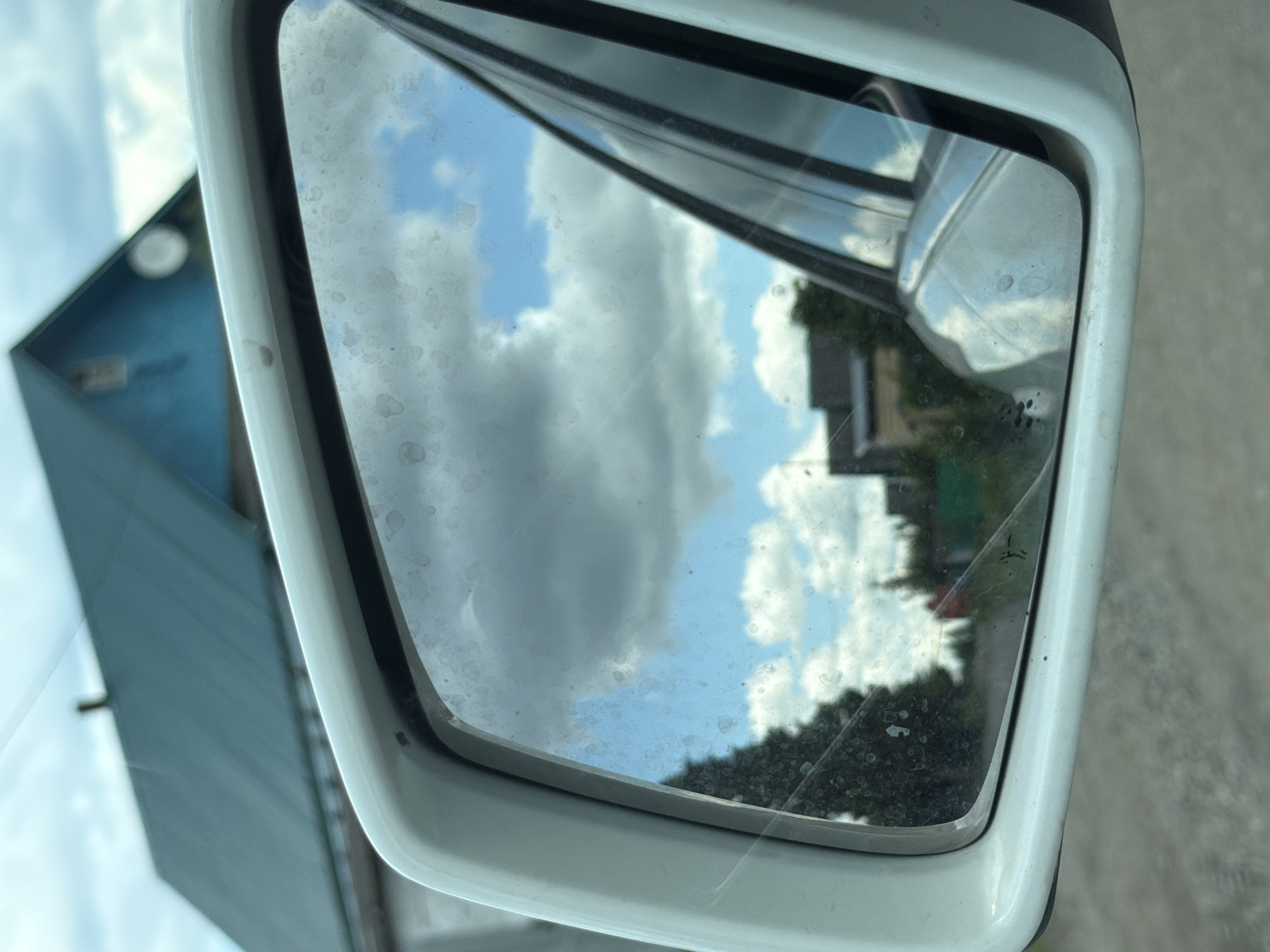 The image shows a reflection in a white car's side mirror. The reflection captures a cloudy blue sky with white clouds, a green roof of a building, and some trees and other structures in the background. The mirror itself appears to be dusty. The setting is likely a rural or suburban area, possibly in Mezhevoy, Russia, given the architectural style of the building. The foreground shows the car's exterior and the road surface.