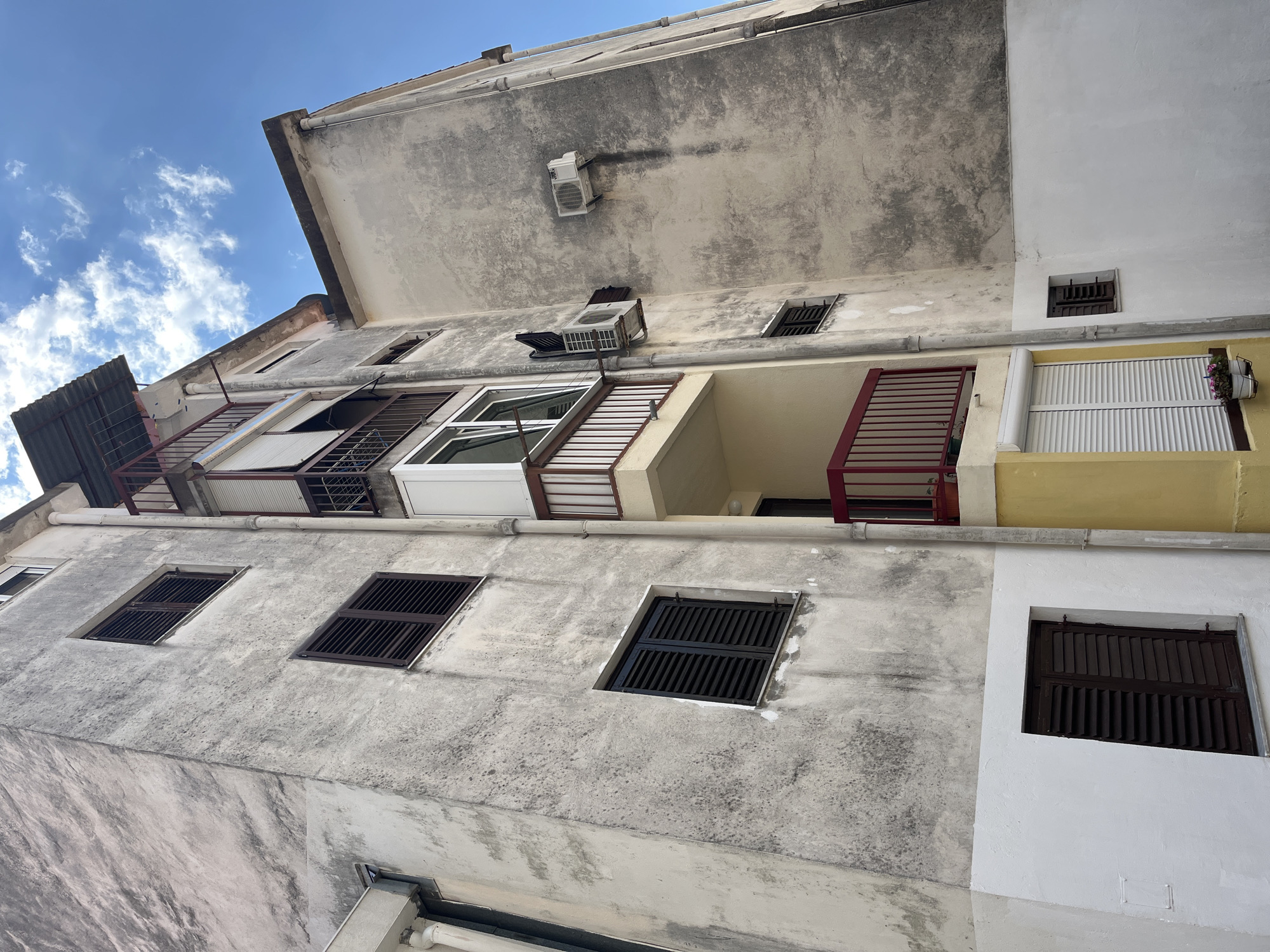The image shows the exterior of an apartment building in Budva, Montenegro, on a sunny day with a blue sky and scattered clouds. The building's facade is weathered and appears to be made of concrete, with patches of discoloration and staining. Several windows with dark brown shutters are visible, as well as balconies with red railings. One balcony has a sliding glass door and a closed roller blind. Two air conditioning units are mounted on the exterior walls. The bottom portion of the building is painted white, contrasting with the upper, older sections. There are no people or animals visible in the image.