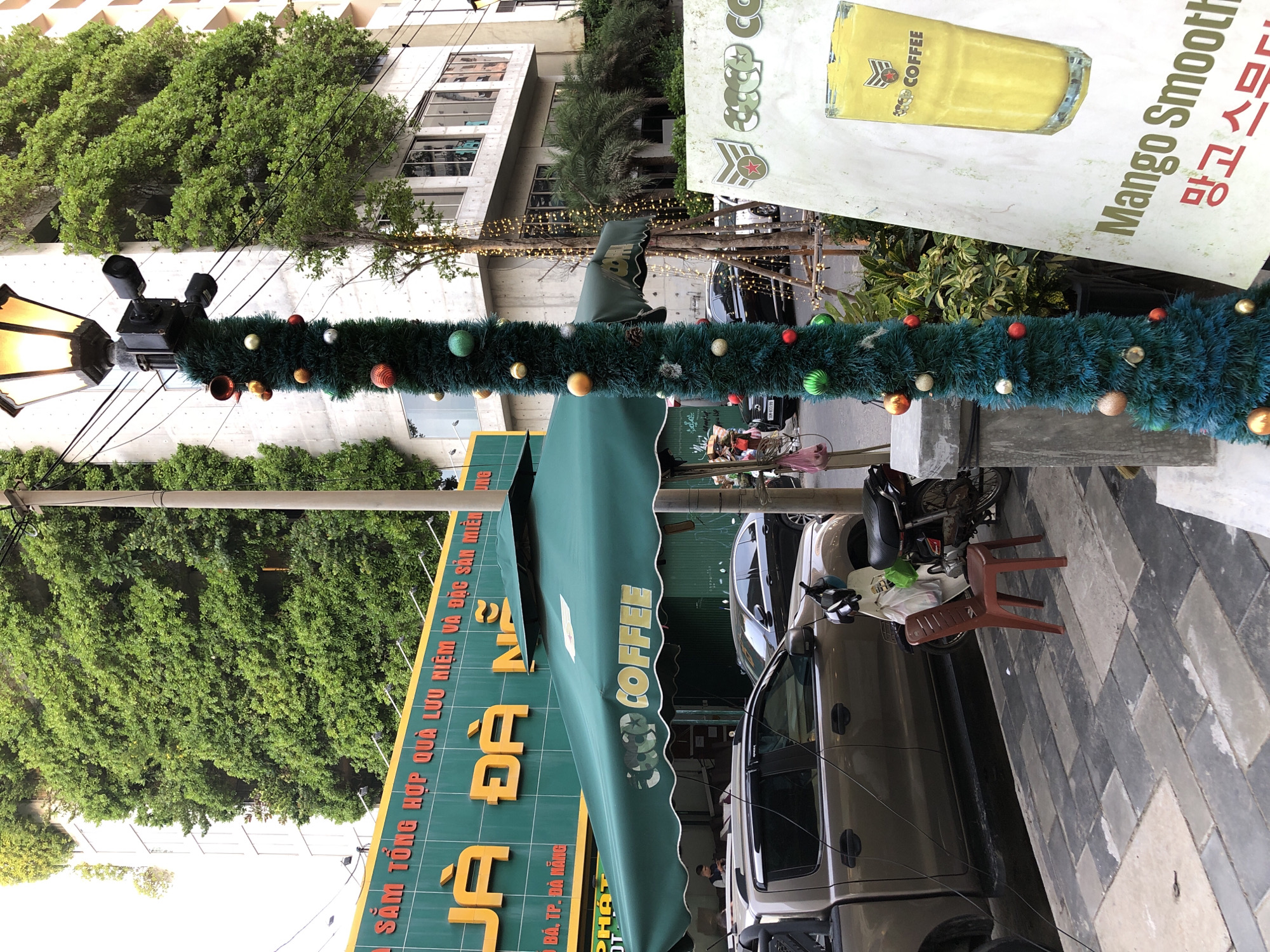 In Đà Nẵng, Vietnam, on a sidewalk, a festive Christmas tree made of green tinsel and decorated with red, gold, and silver baubles is wrapped around a lamppost. The lamppost itself has two security cameras attached to it. Behind the lamppost, a building with lush green vines covering its facade is visible. In front of the building is a green awning with the words "COFFEE" written on it. A pick-up truck and a motorbike are parked on the street, with a single red plastic chair placed on the sidewalk near the motorbike. A sign with a picture of a yellow drink and the words "Mango Smooth" is also visible on the right side of the image. The scene appears to be a street-level view of a coffee shop or a similar establishment, likely decorated for the holiday season.