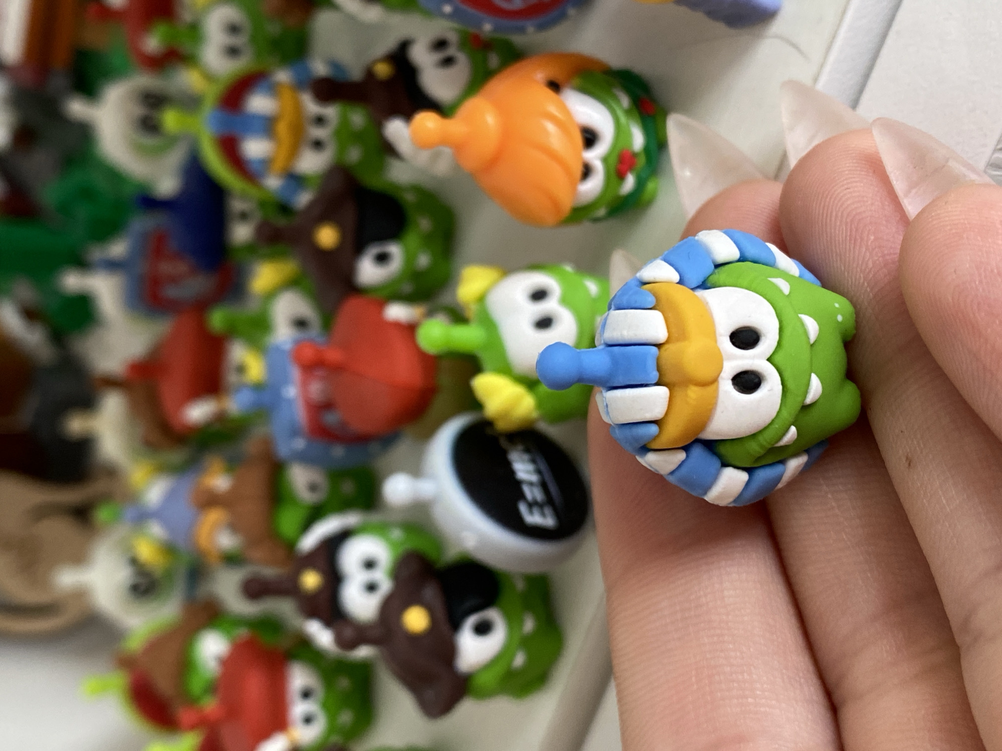 A close-up, shallow depth-of-field shot shows a person's hand holding a collection of small, green, cartoonish figures. The figures have large white eyes with black pupils and wide mouths. Many are wearing different colored hats and accessories. The figure in the foreground is green with white, blue, and yellow striped headwear. In the background, a blurred array of similar figures is visible, some with red, blue, or brown hats. One figure in the middle ground has a gray, circular accessory with "E=mc²" written on it. The overall aesthetic is playful and colorful, suggesting a collection of toys or trinkets. No discernible urban or natural environmental features are present, making location identification within Ostrovki, Russia impossible from the visual information provided.