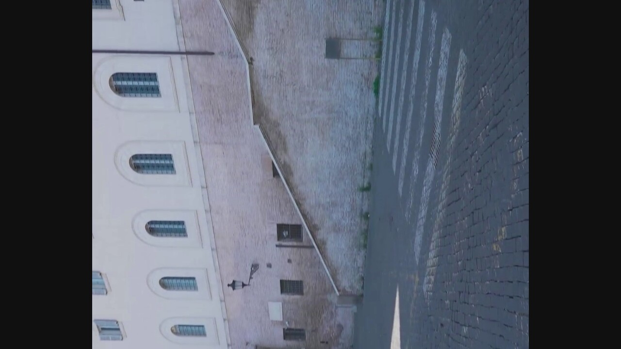 The image captures an outdoor scene in Civita Castellana, Italy. In the background, a light-colored building with arched windows and white trim is visible. A brick wall with several windows and a wall-mounted lamp extends from the building, leading to a set of stairs. The stairs are also constructed of brick. In the foreground, a cobblestone street is shown with a pedestrian crossing. The scene appears to be an architectural setting within the city.