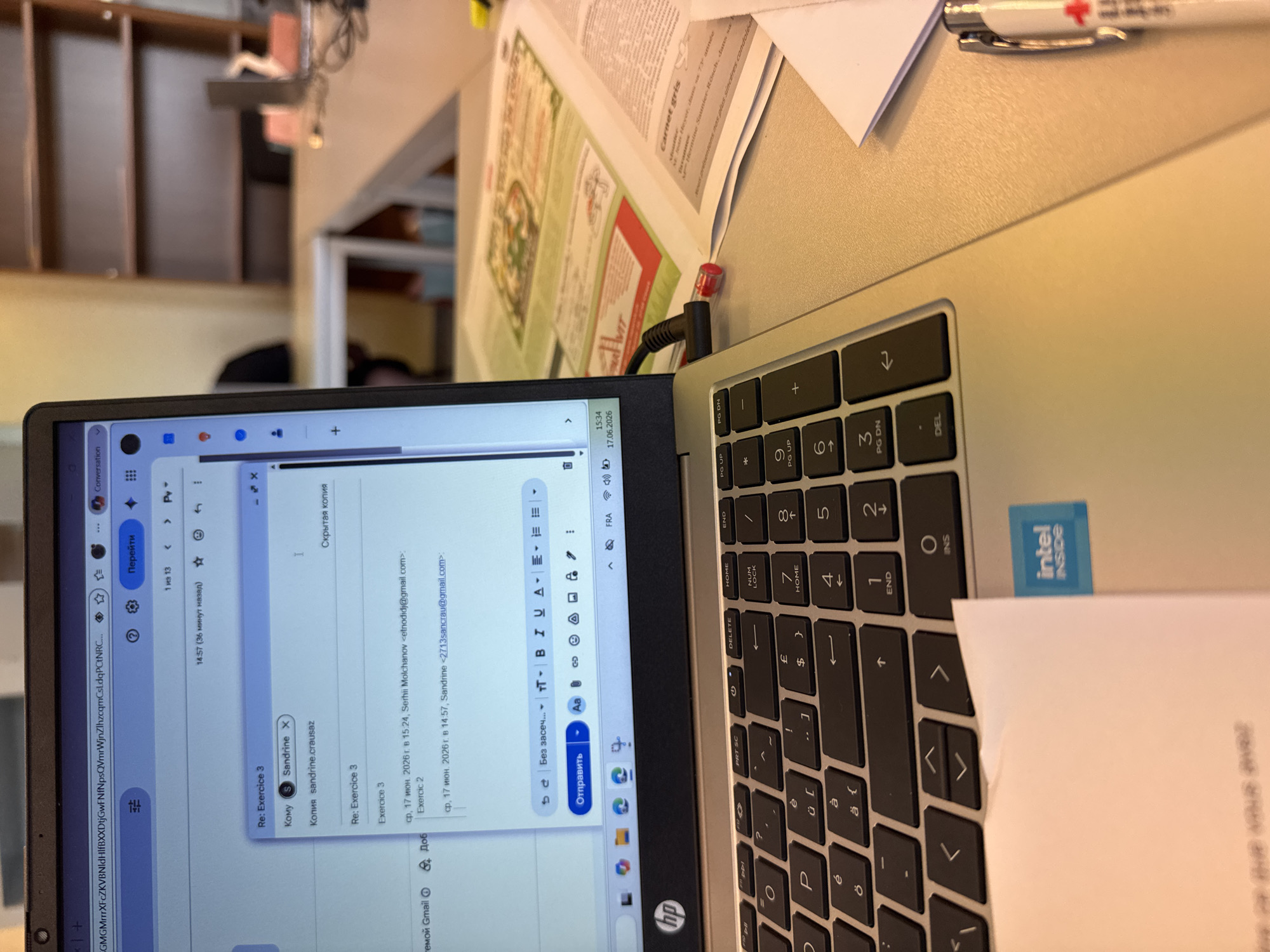 The image shows a laptop with an open email client displaying a reply to an email titled "Re: Exercice 3". The laptop is on a desk, with a keyboard visible. Other items on the desk include papers and a pen. The background shows a blurred view of a room with bookshelves. The scene appears to be an office or study environment. The email client shows sender information, a subject line, and the body of the email. The timestamp on the email indicates it was sent on June 17, 2026, at 14:57. The laptop displays the current time as 15:34 and the date as 17.06.2026. The presence of the "intel inside" sticker suggests the laptop is a Dell or HP brand with an Intel processor. The keyboard has a mix of English and accented characters, indicating a potential European keyboard layout. The environment suggests a typical workspace.