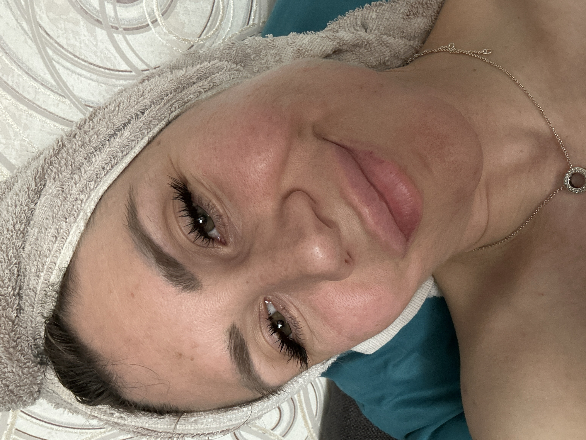 The image shows a close-up, rotated view of a woman lying down with a towel wrapped around her head. She has heavy eyeliner and long, false eyelashes. Her eyes are open, and she is looking towards the camera with a slight smile. A delicate gold chain with a circular pendant is visible around her neck, resting on her chest. The background consists of a teal-colored surface and a white patterned textile. The lighting appears to be artificial, casting soft shadows on her face and neck. There are no visible signs indicating the time of day, weather, or specific location beyond the general context provided. The woman's expression suggests a relaxed or pleased demeanor.