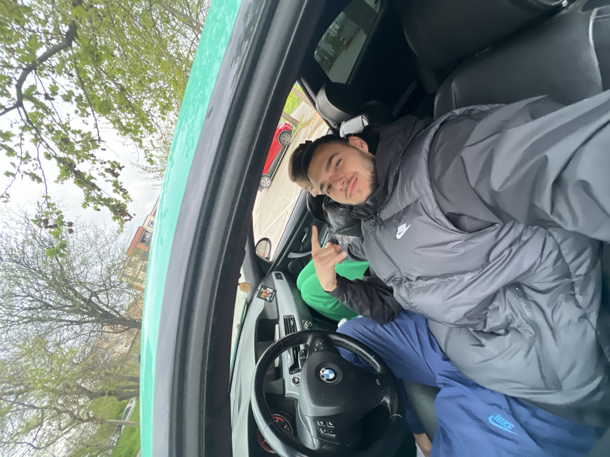The image is a tilted, wide-angle shot taken from inside a green car, likely a selfie or an upward-angled photo. In the foreground, a young man with dark hair and a beard is visible, looking directly at the camera with a slight smile. He is wearing a dark grey puffer jacket with a visible "Nike" logo on the left chest and green pants. His right hand is raised, making a gesture with the index and middle fingers extended. Behind him, part of a second person's head and hair can be seen in the back seat.
The car's interior is dark. A "BMW" logo is clearly visible on the black steering wheel in the lower left quadrant of the frame. Below the main subject, a pair of blue pants with a white "NIKE" logo is visible, likely belonging to him or the other passenger. A small decorative item, possibly a miniature picture frame, hangs from the dashboard or rearview mirror area.
Through the car's windows, an overcast sky is visible, suggesting a cloudy day. Lush green trees, indicative of spring or early summer, frame the upper and lower left portions of the image. In the background, seen through the side windows, a red car is parked, and parts of multi-story buildings, one with orange/red accents, can be observed. The exterior of the car (visible on the left and right edges) appears to be a vibrant green, and there might be water droplets on its surface, suggesting recent rain. The scene portrays a casual moment inside a car, possibly in an urban or suburban setting in Plovdiv, Bulgaria.