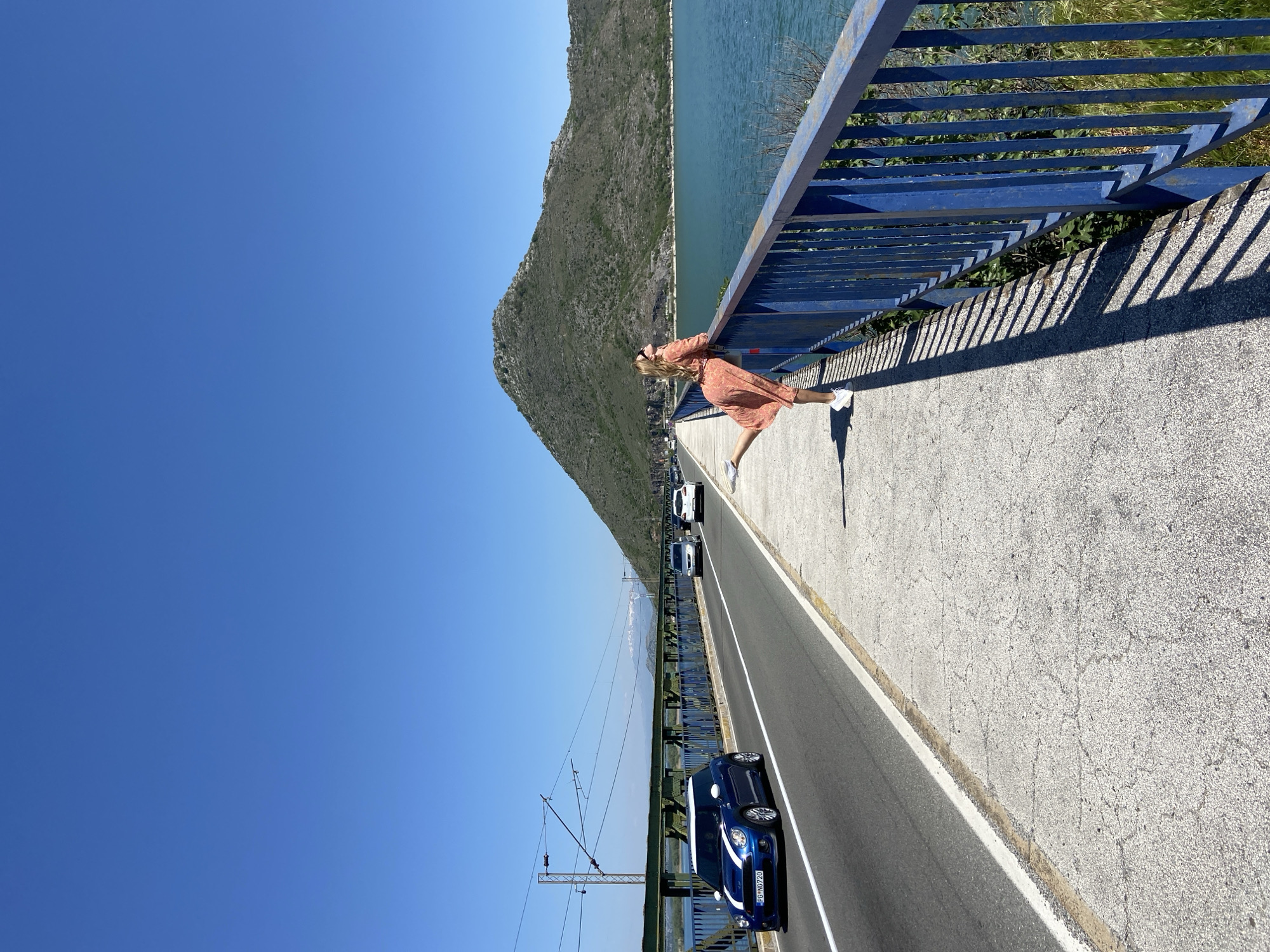 A woman in a peach-colored dress and white shoes is leaning over a blue metal guardrail on a bridge. The bridge has a concrete walkway with a white stripe separating it from a black asphalt road. Cars are driving on the road. The water and a large, rocky, green mountain are behind the guardrail. The sky is clear blue. The bridge is in Kruševica, Montenegro. An overhead power line is on the left.
