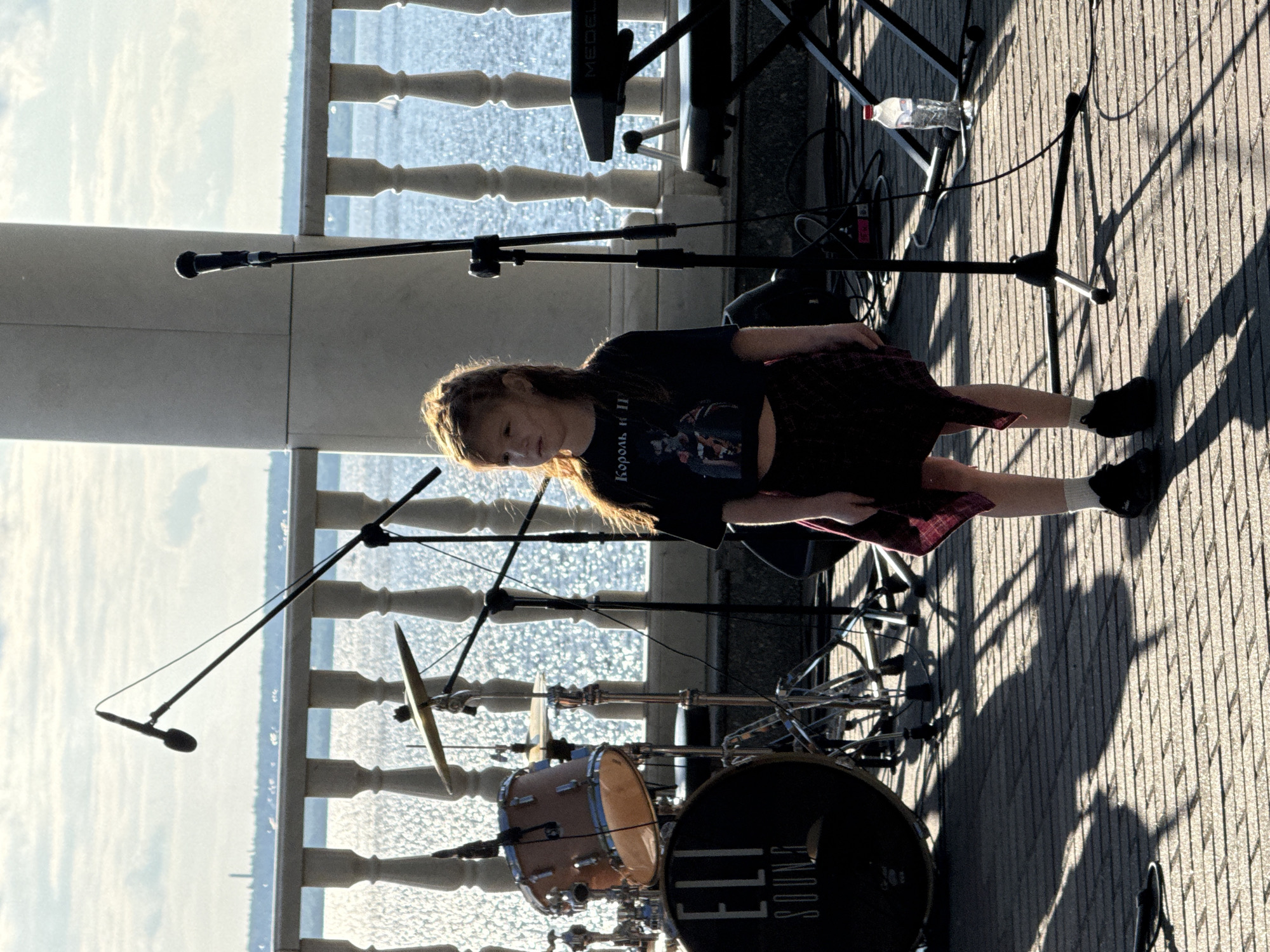 In Postol, Russia, a young girl with long brown hair stands on a paved outdoor stage. She is wearing a dark cropped t-shirt with a graphic print and a plaid skirt. The girl is facing to her left, with her gaze directed towards the water. Behind her, a drum set with the label "ELI SOUND" is visible, along with microphones and musical equipment. A keyboard is positioned to the right. The scene is set against a backdrop of a bright, sunlit body of water, with a railing and distant structures suggesting a waterfront promenade or park. The sunlight casts long shadows across the stage.