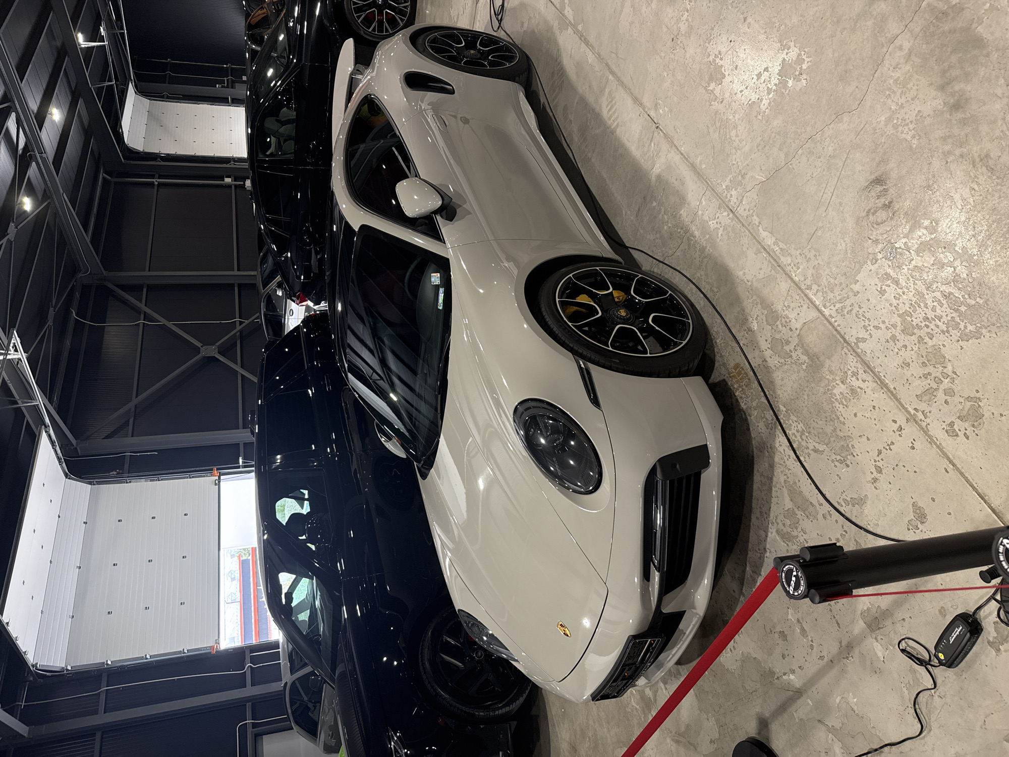 The image shows a light gray Porsche 911 parked indoors, likely in a showroom or garage. A black vehicle is parked next to it. A black barrier with a red rope is positioned in front of the Porsche. The floor is concrete with visible imperfections. The background features a black industrial-style wall and a white door or panel. The location is possibly in Plovdiv, Bulgaria, but no definitive evidence confirms this.