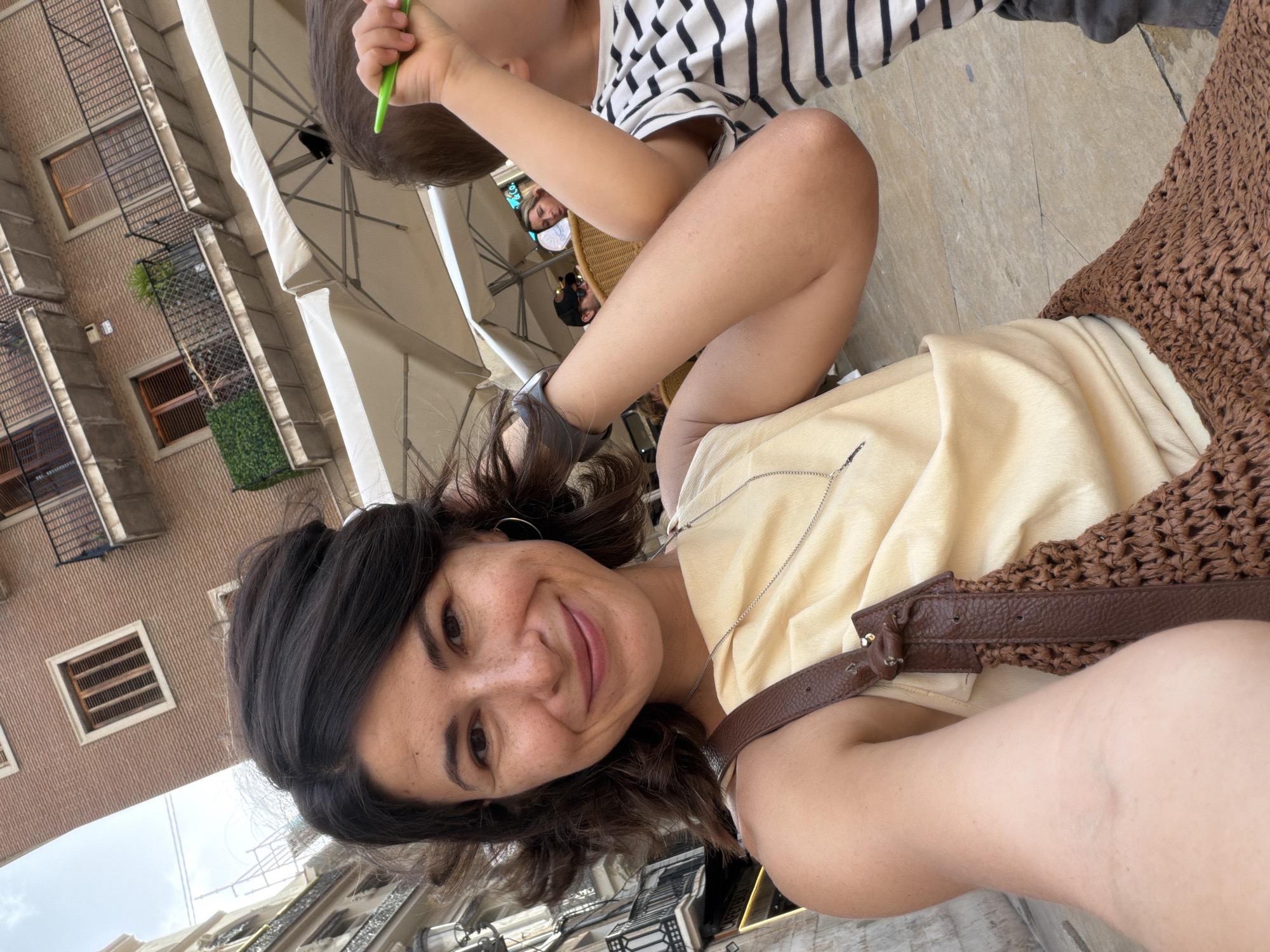 An outdoor cafe scene is captured in an eye-level, close-up selfie with a smiling woman in the foreground. She has dark, wavy hair and is wearing a light yellow sleeveless top with a brown woven bag draped over her shoulder. A delicate silver necklace adorns her neck. To her right, a child in a black and white striped t-shirt is partially visible, holding a green object. In the background, an awning of a cafe is visible, along with the facade of a building with balconies and windows. Other patrons and cafe umbrellas are also present in the background, suggesting a public dining setting in an urban environment. The floor appears to be paved with large square tiles.