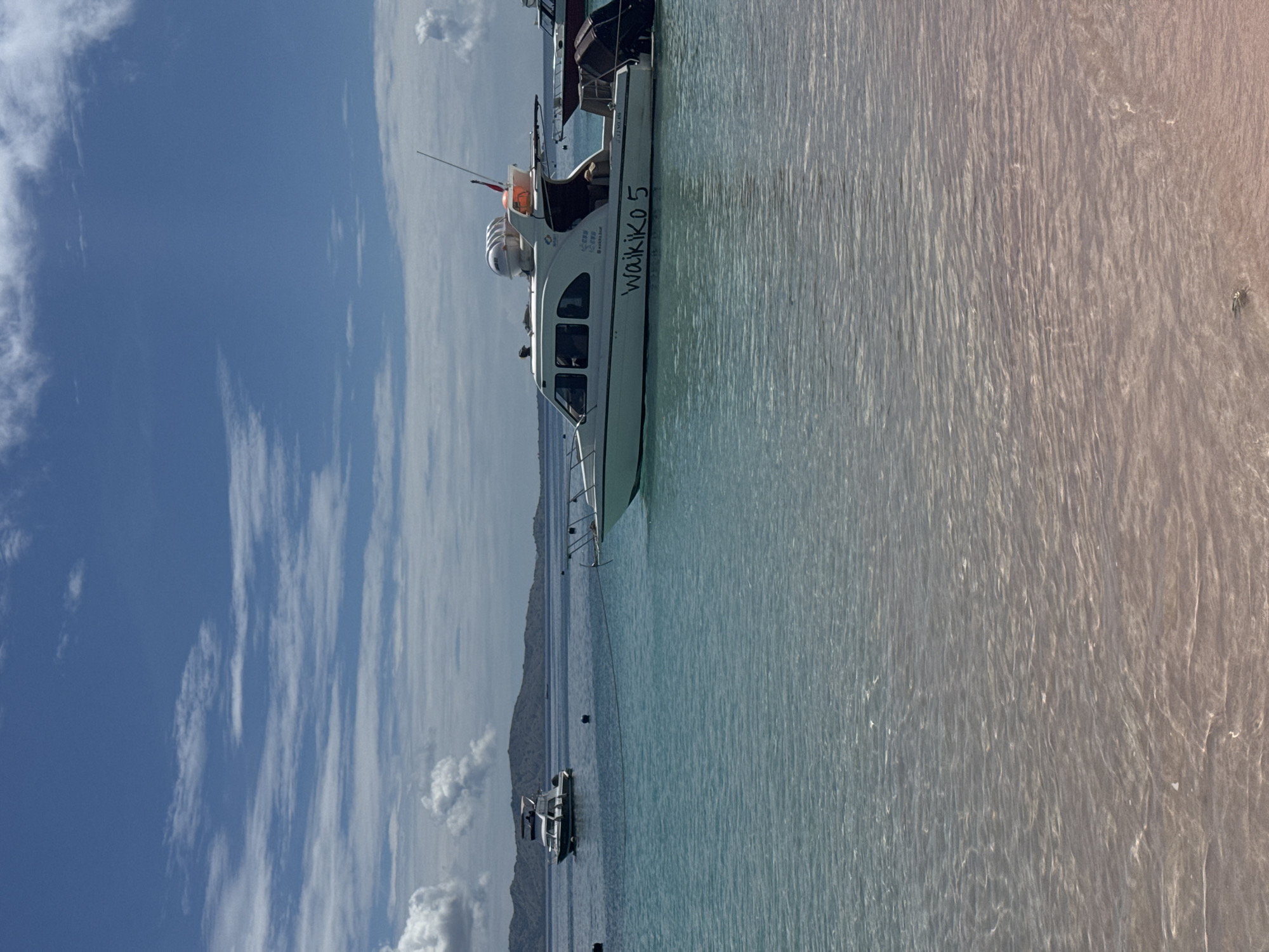 A white boat named "Waikiko 5" is prominently featured, floating on calm, clear water under a bright blue sky with scattered white clouds. The boat, which appears to be a passenger or tour vessel, has multiple windows along its side and a small antenna or mast at its stern. On its side, above the name, smaller text is visible, including "CAP. 20 PERS" (Capacity 20 Persons) and "WAIRIKI BOAT".
In the background, a distant shoreline or mountain range stretches across the horizon. Another, smaller white boat is anchored or moored further out in the water. The water itself is a clear, light blue or turquoise near the foreground, transitioning to a deeper blue further away, and shows subtle ripples on its surface, reflecting the sunlight. A faint gradient of warmer, reddish-orange light is present on the far right side of the water, possibly due to natural light conditions or reflections. The scene suggests a peaceful daytime setting, likely in a coastal or large lake environment with good weather.