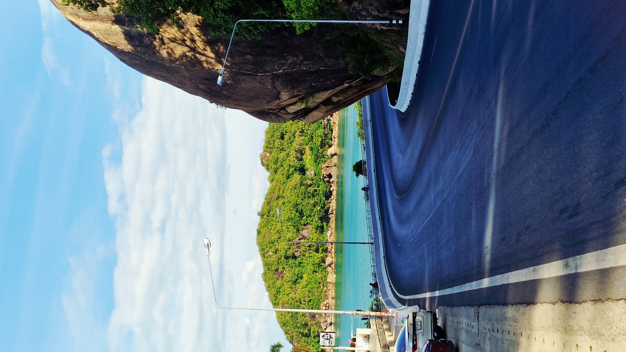 The image depicts a scenic coastal road in Baan Chaweng Noi, Thailand. A dark asphalt road with white lane markings curves through the frame, bordered by a white concrete barrier on the right and a sidewalk with parked cars on the left. To the left of the road, a turquoise body of water stretches towards a lush green, tree-covered island with rocky outcrops. The sky above is a bright blue with scattered white clouds. A large, dark boulder dominates the right side of the image, casting shadows across the road and a nearby tree. Two streetlights are visible along the road. On the sidewalk, a few people are present, and several cars are parked. The scene captures a beautiful tropical landscape with a clear view of the ocean and the island.