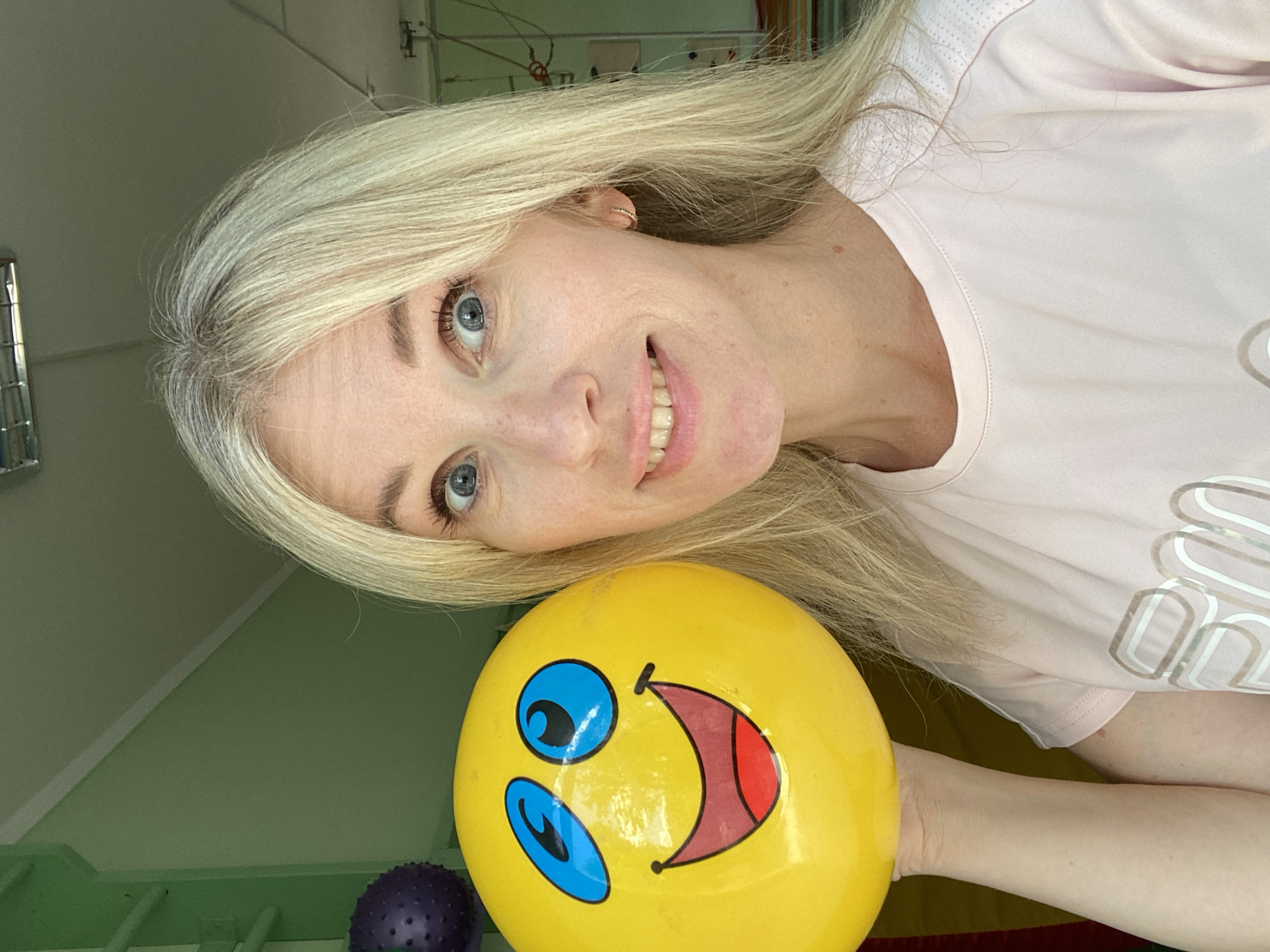 A blonde woman with blue eyes and fair skin is pictured in a selfie, holding a large yellow ball with a smiley face printed on it. She is wearing a light pink t-shirt. The background appears to be an indoor space with green walls, and some exercise equipment, including a purple spiky ball and a ladder-like structure, are visible. The overall environment suggests a gym or a recreational area. The woman's expression is one of amusement or happiness, with a wide smile and bright eyes.