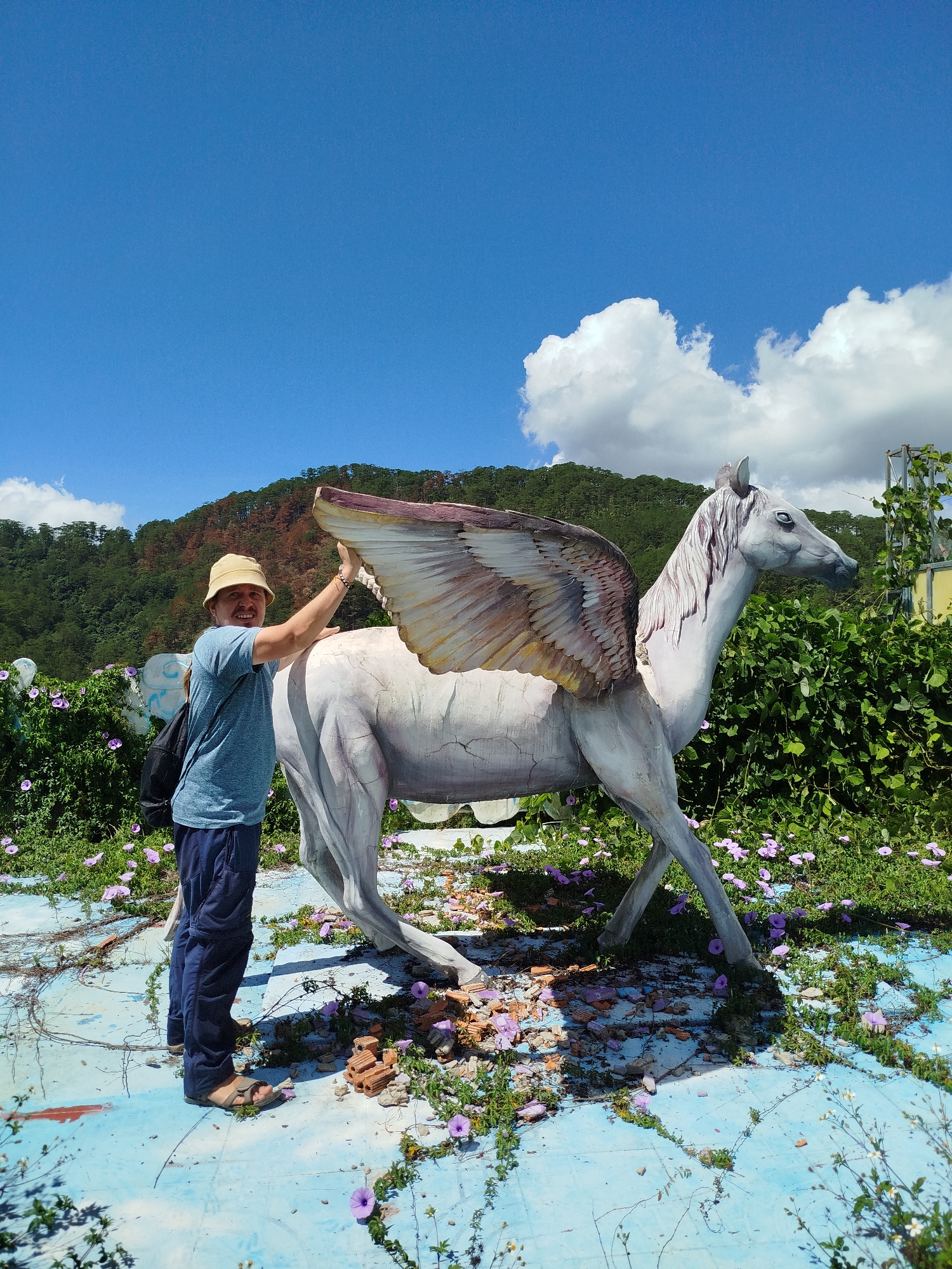 A man in a khaki hat, blue shirt, and blue pants stands next to a white, winged horse statue outdoors. The statue is cracked and weathered, surrounded by greenery and purple flowers on a light blue surface that appears to be damaged. Behind them, a wooded hillside rises under a bright blue sky with scattered clouds. This image appears to be taken in the city of Da Lat, Vietnam.