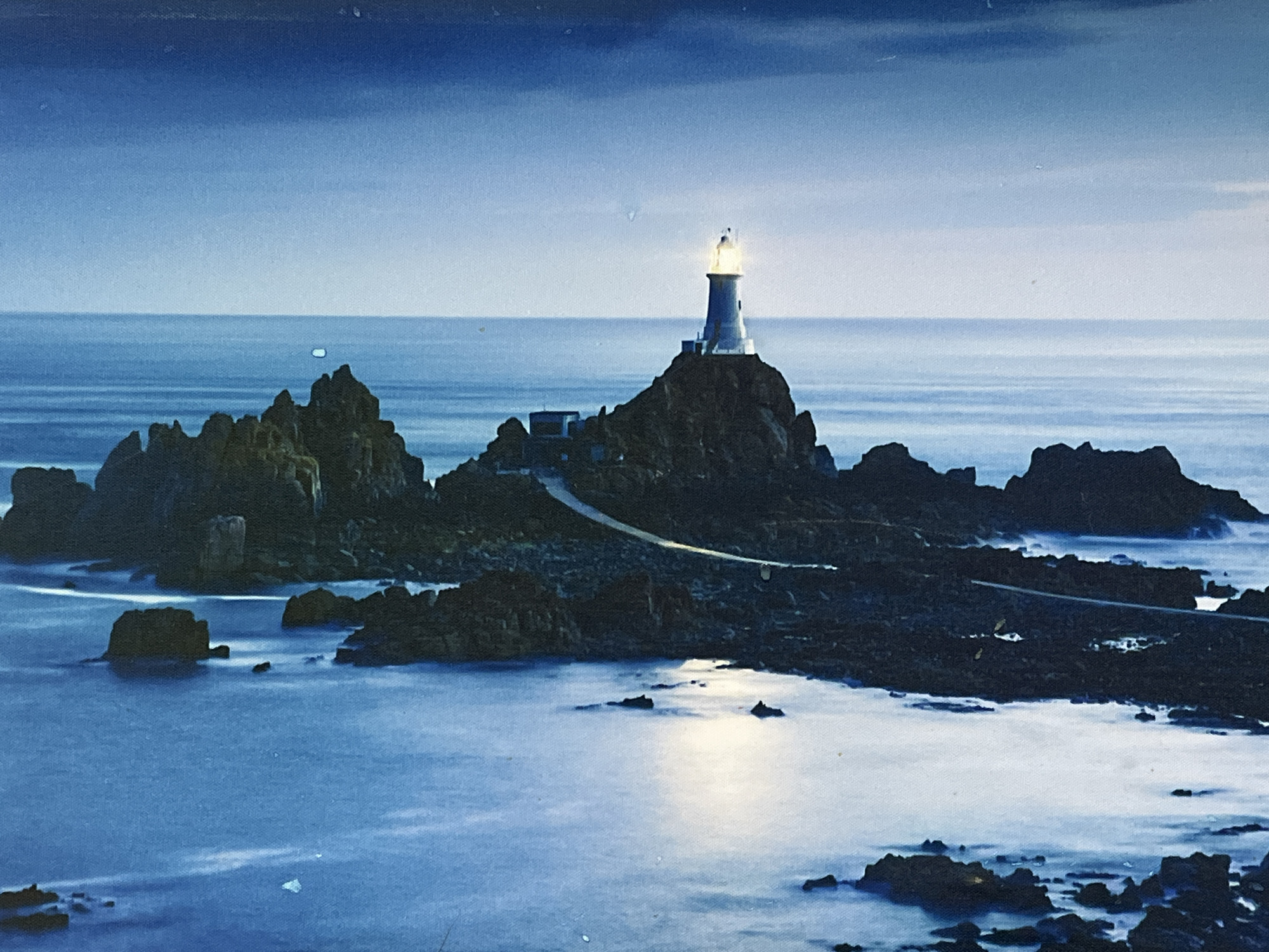 A lighthouse stands on a rocky island surrounded by calm ocean waters. The sky is a deep blue, fading to a lighter blue towards the horizon. The lighthouse itself is white with a glowing lantern at its top, casting a beam of light. A pathway made of stones winds its way up the rocky island towards the lighthouse. The rocks are dark and jagged, with some of them partially submerged in the water. The water is a deep blue, with streaks of white indicating gentle movement. The scene is peaceful and serene, with a sense of isolation. The image captures a twilight or early evening setting.