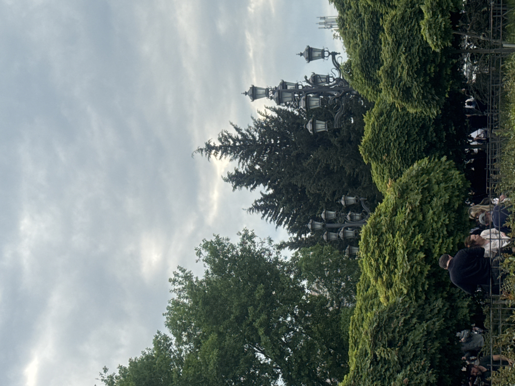 The image shows a street scene in Tbilisi, Georgia. Several trees and bushes are visible, including a large pine tree. There are ornate street lamps. A group of people stand or sit behind a metal fence. The sky is overcast with light gray clouds.