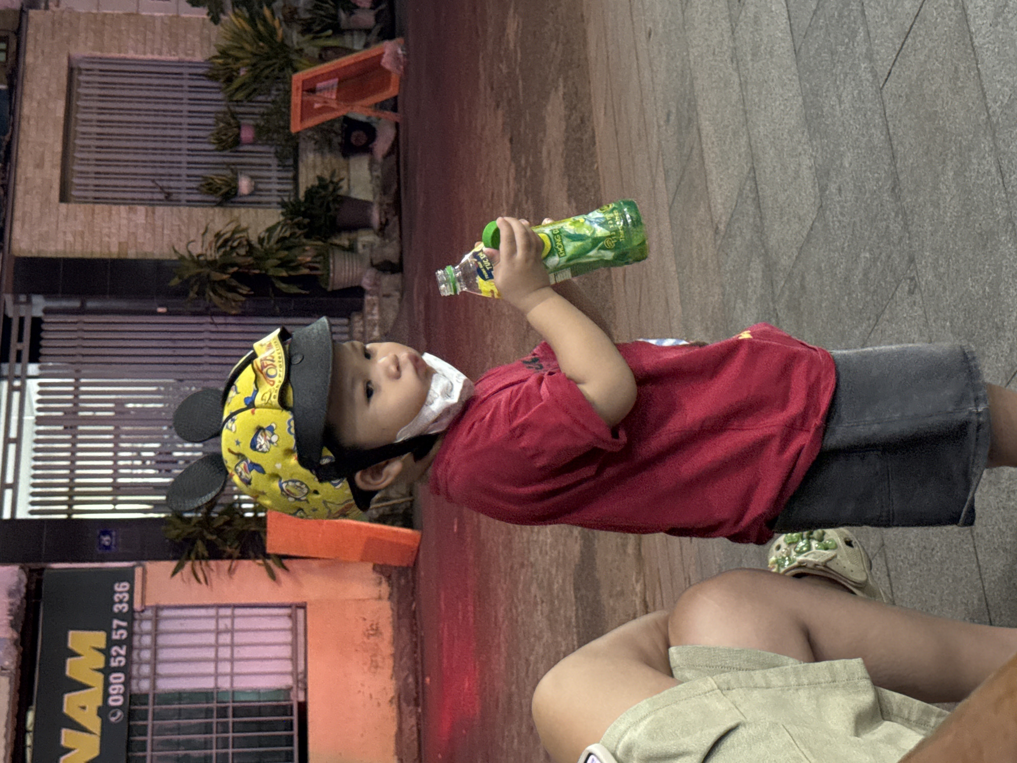 The image shows a young child wearing a yellow helmet with Mickey Mouse ears and a red shirt. The child is holding a green bottle of a beverage. The child is standing on a sidewalk in front of a building with a sign that reads "NAM" and a phone number. The background includes potted plants, a metal gate, and a red display stand. The scene appears to be at night, with artificial lighting illuminating the area. The context of Nha Trang, Vietnam is provided by the sign and the overall setting, suggesting an urban street scene.