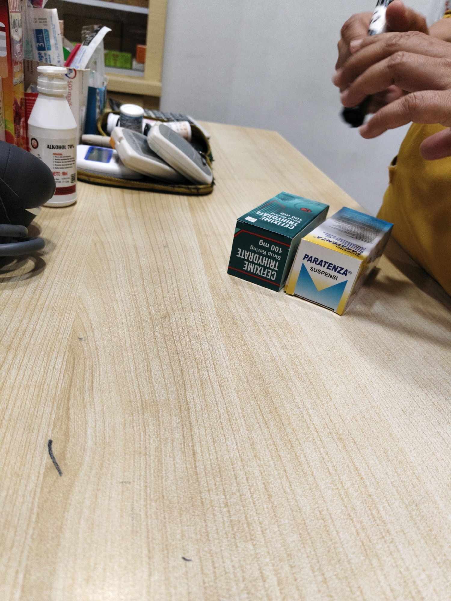 In Lhokseumawe, Indonesia, on a light-colored wooden table, there are several medical items. On the left, a white bottle labeled "ALKOHOL 70%" stands near a dark gray object. Behind it, a medical bag is open, containing a glucose meter and other items. Two small boxes of medicine are prominent in the foreground. The box on the left is green and white, with text that includes "CEFIXIME TRIHYDRATE" and "100 mg". The box on the right is blue and yellow, labeled "PARATENZA" and "SUSPENSI". In the upper right corner, a person's hands are visible, holding a black and silver pen. The background is blurred, showing shelves with more items.