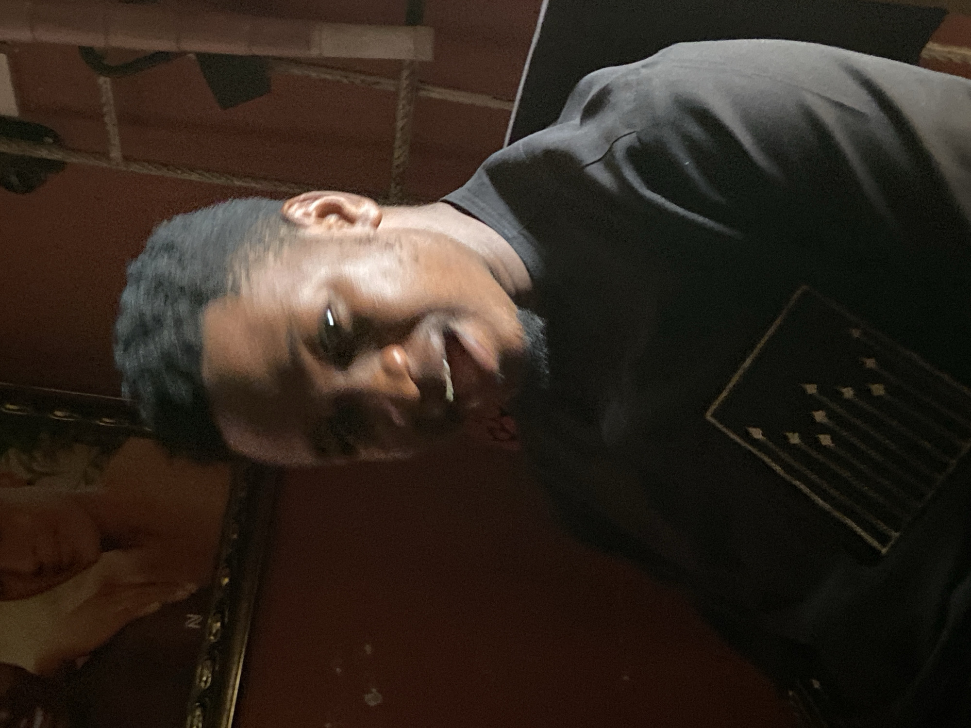 An indoor, dimly lit scene in Mararaba, Nigeria, features a young Black male in the foreground, facing slightly right and looking towards the viewer's left. He has short, dark, wavy hair and light facial stubble around his jawline and chin. His mouth is open, revealing white teeth, suggesting an animated expression, possibly speaking or reacting. He is wearing a black, short-sleeved crew-neck shirt with a light-colored geometric design on the chest, composed of multiple parallel vertical lines topped with small diamond or arrow-like shapes.
The background consists of dark reddish-brown walls. To the left of the subject, an ornate gold-colored picture frame is mounted on the wall, containing a faded portrait of a light-skinned individual, appearing to be a woman with bare shoulders, with the letter "N" faintly visible below the image. To the right of the subject, a ladder-like structure, seemingly constructed from dark cord or rope with horizontal rungs, leans against the wall. A dark, unidentified object is suspended from this structure. The lighting is low, casting prominent shadows and highlighting the subject's face from a front or front-left source.