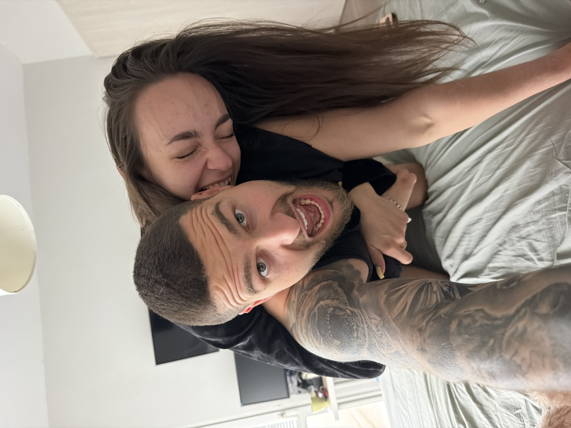 A horizontally oriented, high-angle selfie depicts a man and a woman in close proximity. The man, positioned at the bottom of the frame and facing the camera, has short brown hair, a beard, and visible tattoos on his right arm. His mouth is open in a wide expression. Above him, the woman is biting his head. She has long brown hair that is spread out and a closed-eyed expression. They are both wearing black shirts.
The background consists of a gray bed, a white wall, and a black television screen. The location is unknown and the prompt to include Sofia, Bulgaria should be disregarded as it is not possible to determine the location from the image.