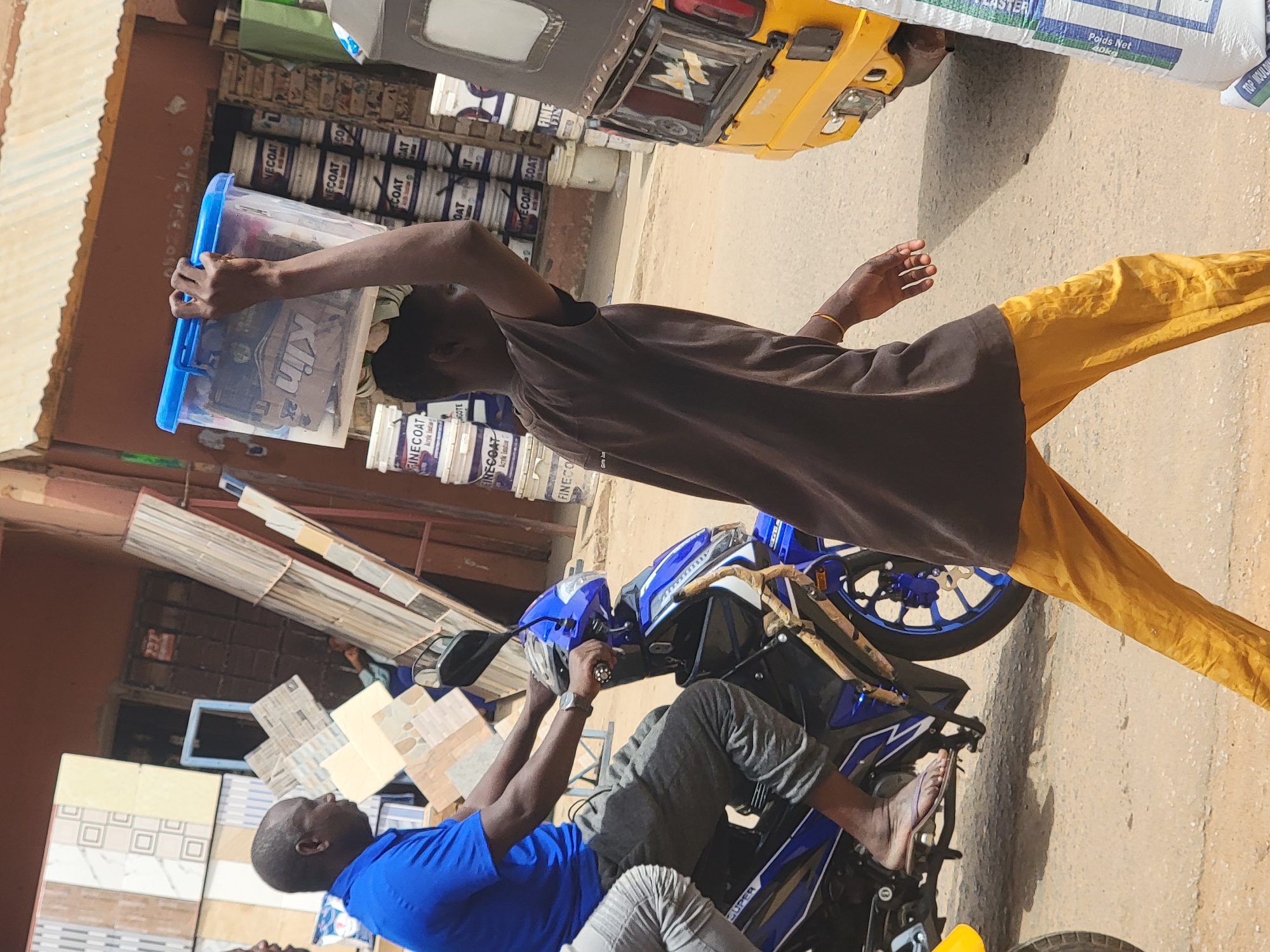 Here's a detailed description of the image:
**Location:** The image appears to be taken on a street in Maiburiji, Nigeria, as indicated by the name on the plaster bag in the upper right corner of the image.
**People:**
* A man wearing a dark brown shirt and yellow pants carries a blue plastic container on his head.
* Another man, wearing a blue shirt and gray pants, is sitting on a blue and white motorcycle. He appears to be adjusting something near the motorcycle's handlebars. He is barefoot.
**Objects:**
* Blue plastic container.
* Blue and white motorcycle.
* Various tiles and materials are stacked and displayed near the man on the motorcycle.
* Cans and containers, possibly of paint, are stacked behind the man carrying the blue container.
* A three-wheeled vehicle, yellow in color, is partially visible at the top of the image.
* A white bag is sitting on top of the three-wheeled vehicle. It has the words "TOP MOULD" and "Poids Net 40kg" on it.
**Actions:**
* The man in the brown shirt is walking with a container on his head.
* The man in the blue shirt is working on the motorcycle.
**Environment:**
* The setting appears to be an outdoor marketplace or a roadside business area.
* The ground is paved and dusty.
* There are various building materials and goods visible in the background.