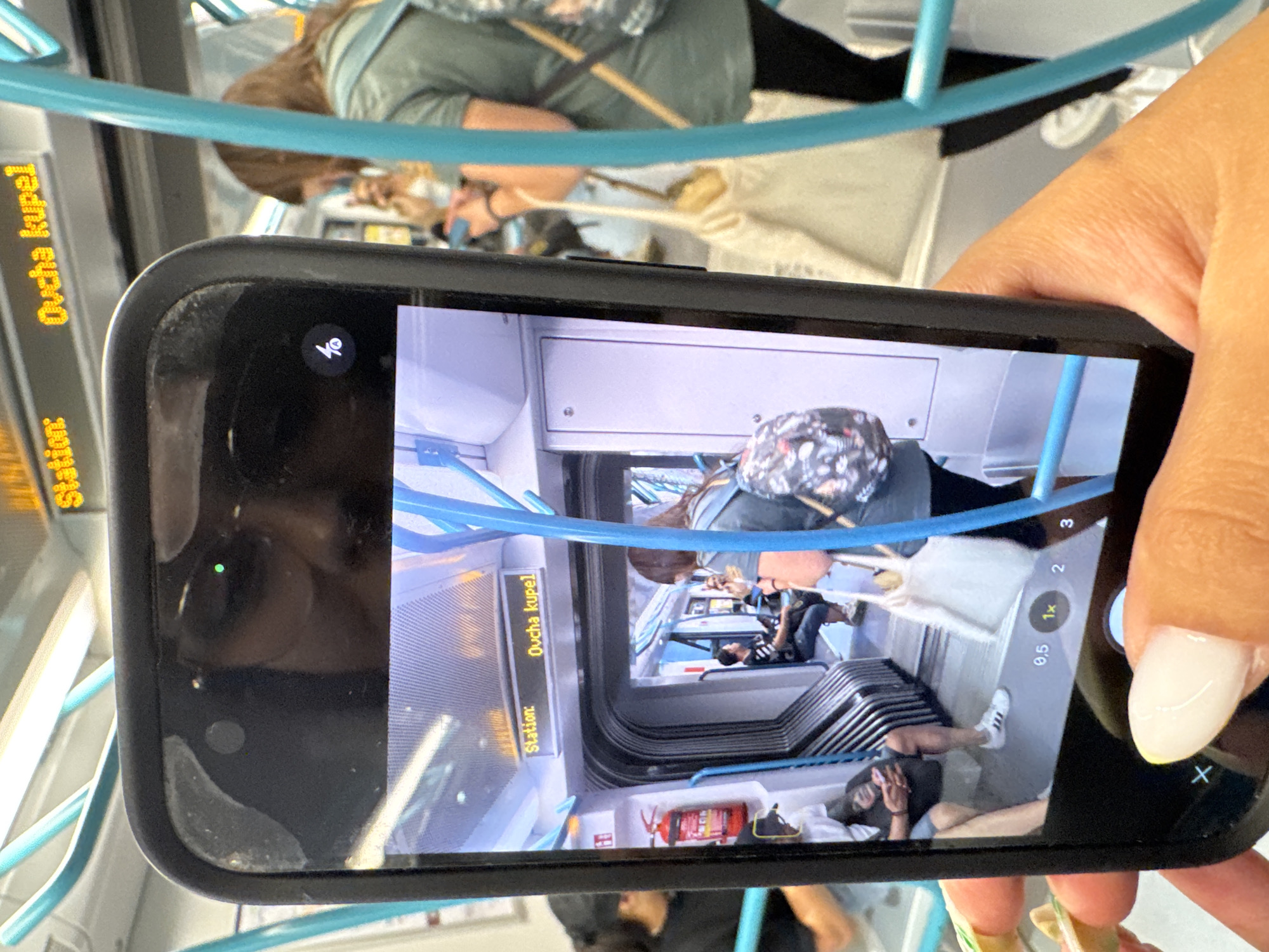 The image shows a person holding a smartphone, capturing a scene inside a public transportation vehicle, likely a metro train, given the interior design and overhead signage. The train appears to be at a station, as indicated by the digital display that reads "Station: Oucha kupel". This station name is located in Sofia, Bulgaria.
Inside the train, there are several passengers. In the foreground of the captured image, a person with a backpack is visible, standing near a blue pole. Further inside the train, a person is seated, facing away from the camera. On the right side of the captured image, another passenger with a bag is seen. The interior of the train features prominent blue poles for support and a metallic-looking seating area. A red fire extinguisher is mounted on the wall on the left side of the captured image.
The smartphone itself is in the foreground of the primary image, with the screen displaying the captured scene. The phone's camera interface is partially visible at the bottom of the screen, showing zoom options (0.5x, 1x, 2, 3) and a shutter button, with a finger about to press it. The phone's reflection is also visible in the top part of its own screen, showing the photographer's face partially obscured by their glasses.