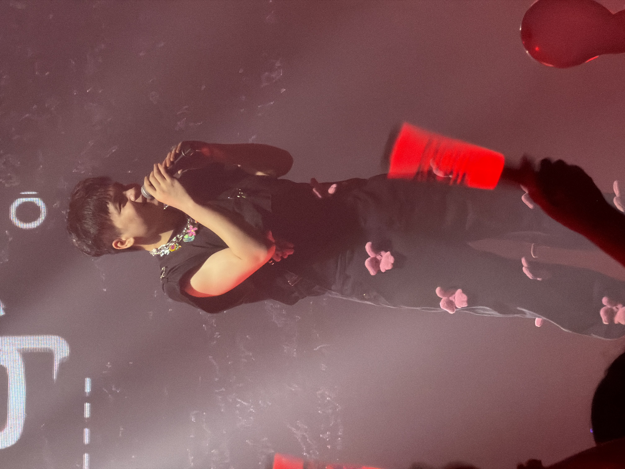 The scene is a live performance in 江北城街道, China, featuring a male performer on a stage bathed in reddish ambient light. The performer, an East Asian male with short dark hair, stands center-frame, holding a dark microphone to his mouth with both hands while smiling. He wears a black sleeveless top accented by a colorful, multi-component necklace or collar embellishment featuring various bright hues (green, pink, yellow, blue, purple). His dark trousers are adorned with numerous small, three-dimensional, pink plush objects, resembling rabbit heads or cartoon characters, distributed across both legs. The background is dark and hazy, with visible white luminous segments of text or graphics, including a prominent capital "O" and a partial "J" on an overhead screen or backdrop. In the lower foreground, hands from an unseen audience clutch bright red, elongated light sticks, and a large, round red object, possibly a balloon, is visible on the far right.