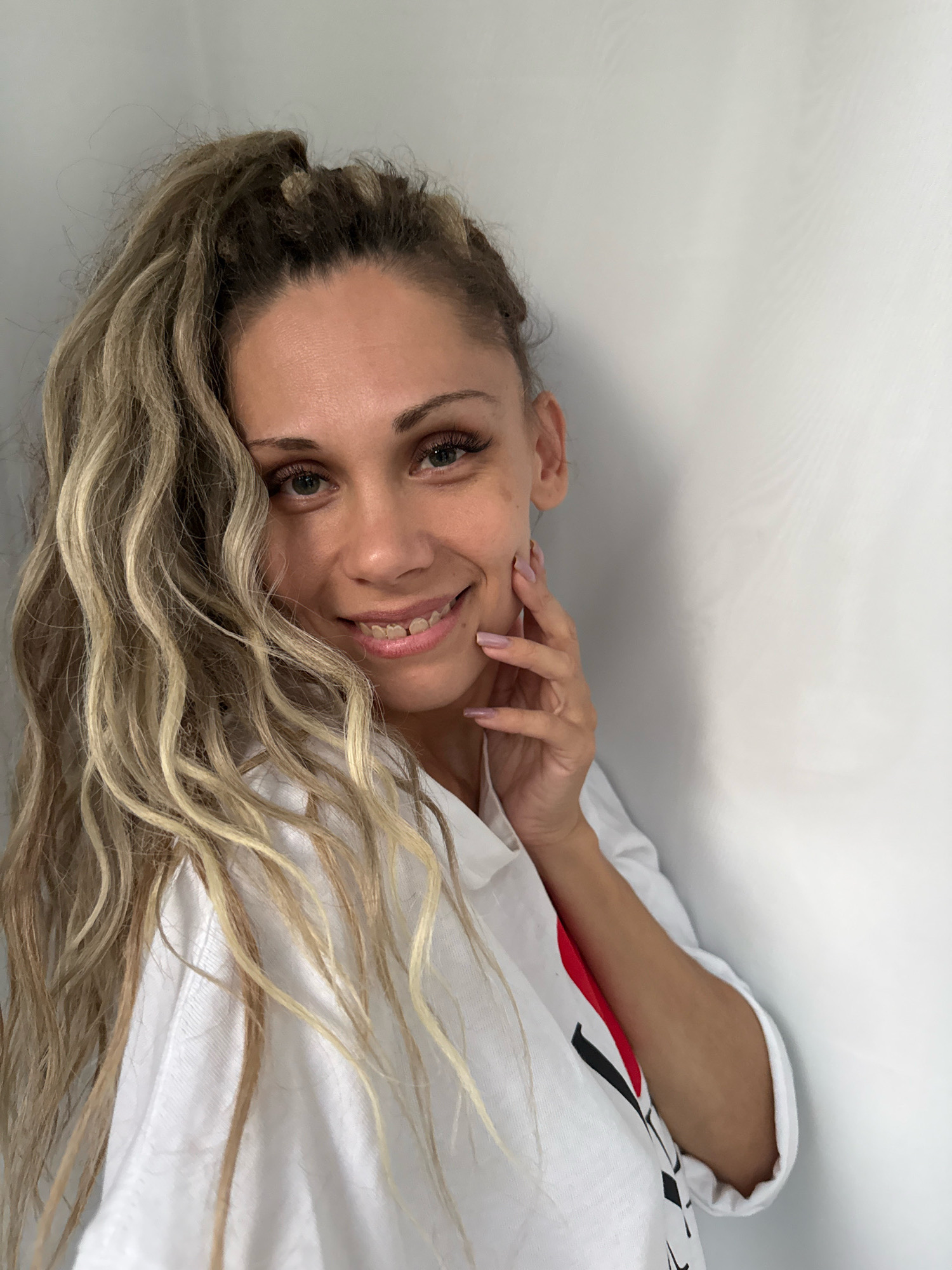 The image shows a woman with blonde, wavy hair pulled up in a high ponytail. She is wearing a white shirt with a red and black design on it. Her left hand is touching her cheek. She is smiling. The background is a plain white wall. The image was taken in Rusanovo, Russia.