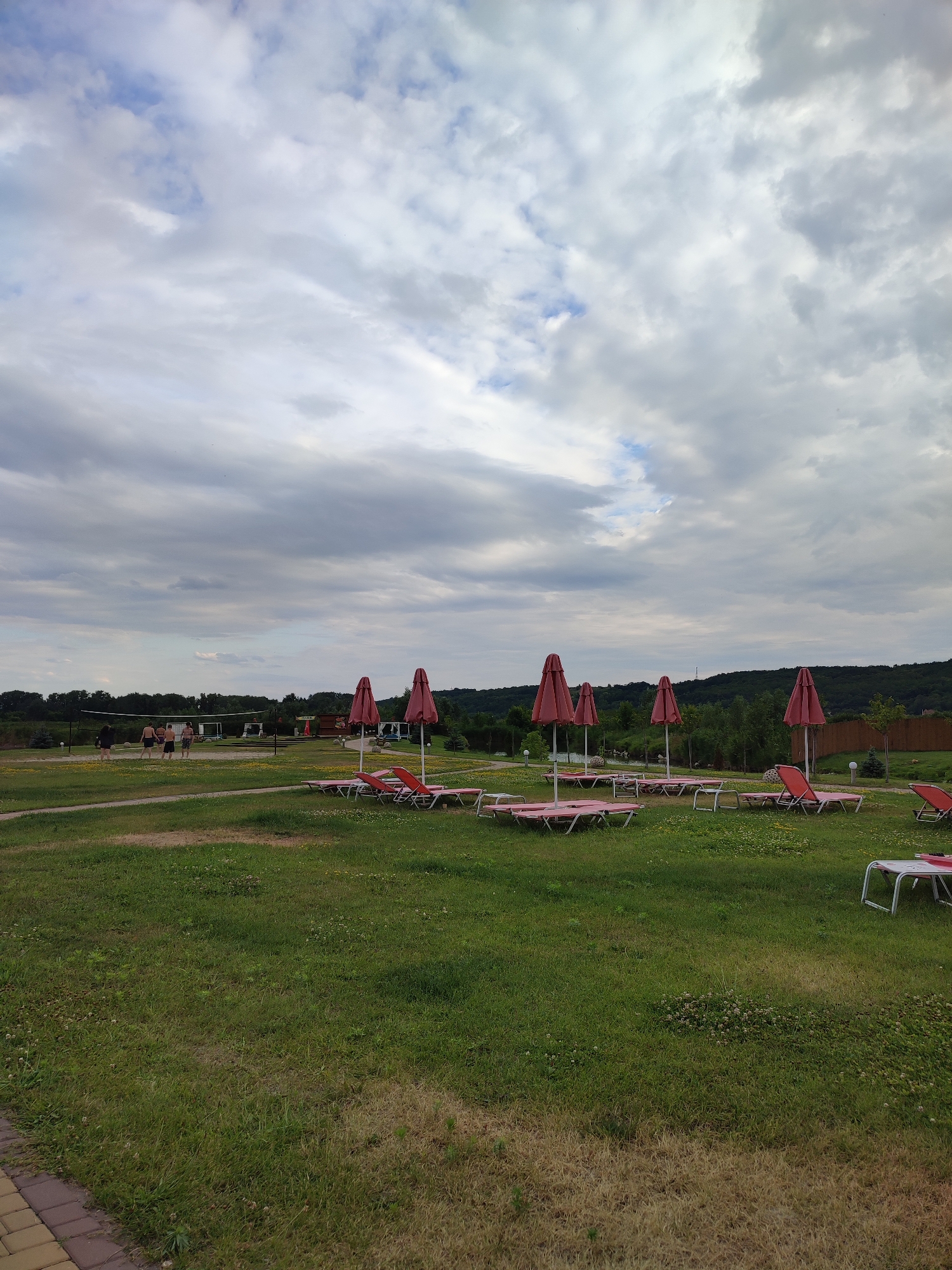 The image depicts an outdoor scene in Shcherbani, Ukraine, likely a recreational area. In the foreground, a well-maintained lawn is dotted with small white flowers. A paved walkway with a yellowish-brown border runs along the bottom left edge. Scattered across the lawn are several red lounge chairs with white frames, some of which are occupied by folded pink cushions. Several closed red umbrellas are positioned behind the lounge chairs, some on their own stands and others associated with the chairs.
In the middle ground, a group of five individuals, appearing to be men and women, are engaged in a game of volleyball on a sand court. A net is set up for the game, with a structure resembling a small kiosk or building in the background. Further in the background, a line of trees forms the horizon, with a gentle slope rising behind them. The sky is overcast with a mixture of white and gray clouds, suggesting a moderate to cloudy day. The overall atmosphere is one of a peaceful, perhaps slightly subdued, day at a park or resort.
