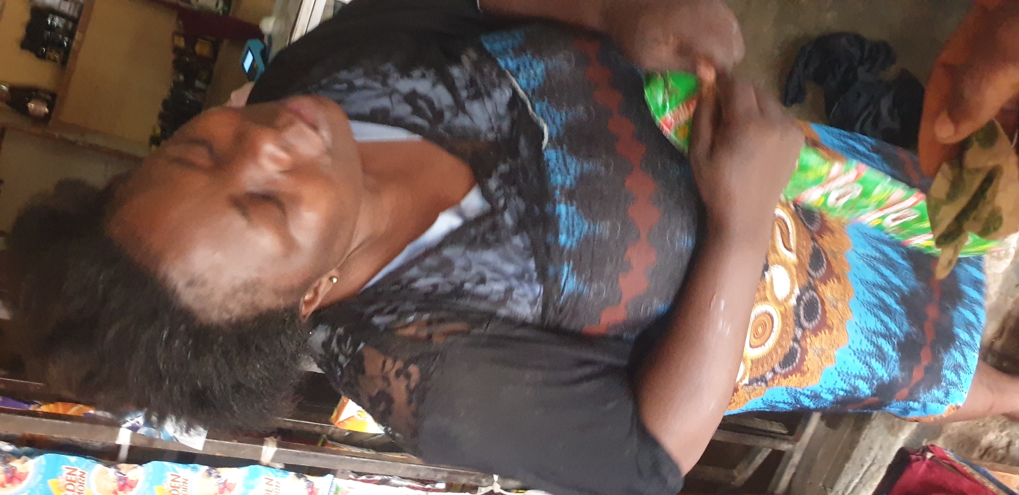 A medium shot captures a woman in Sabon Tasha, Nigeria, appearing to be in a retail setting. The woman, positioned in the center of the frame, has dark, kinky hair styled upwards and is wearing a black lace top over a patterned garment featuring blue and brown designs. She is holding a green and white packaged item in her hands, with a portion of a blue and orange patterned fabric visible below her waist. To her left, several colorful packages are stacked vertically, suggesting they are merchandise. In the background, shelves are stocked with various bottles and products, indicative of a shop. The lighting is somewhat dim, and the overall scene suggests a casual, everyday moment.