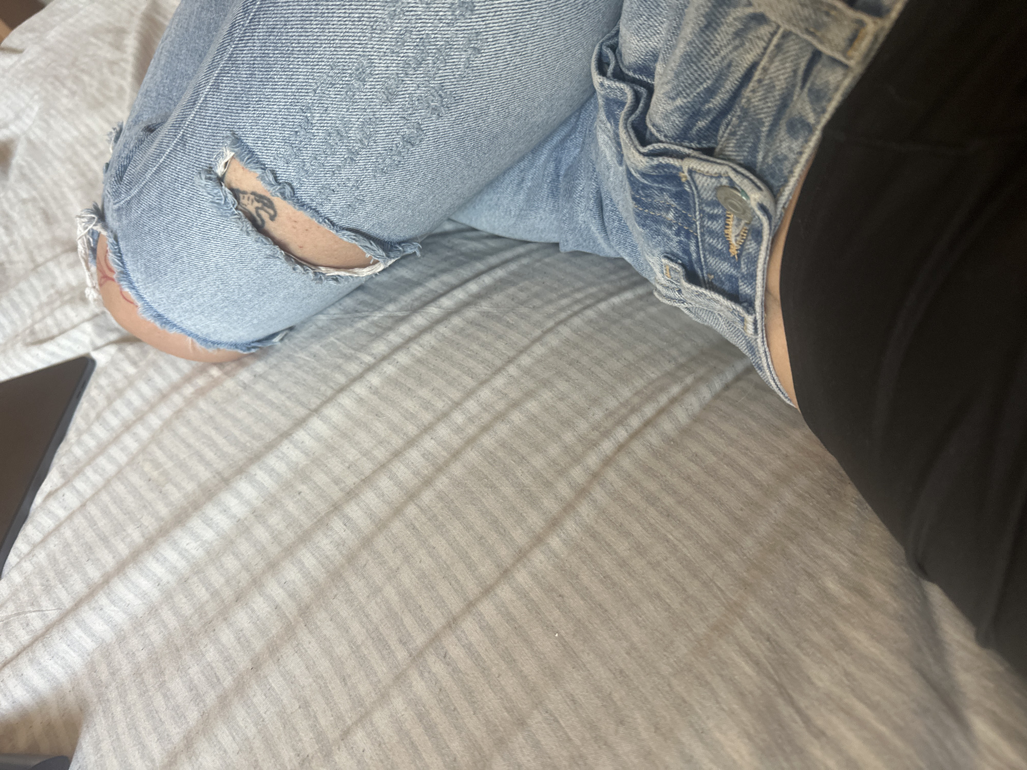 The image depicts a person's leg on a striped bedsheet. The person is wearing light blue ripped jeans and black leggings. A small tattoo is visible on the leg where the jeans are ripped. A corner of a laptop is seen on the lower left. The setting does not appear to be in the city of Sirakovo, Bulgaria.