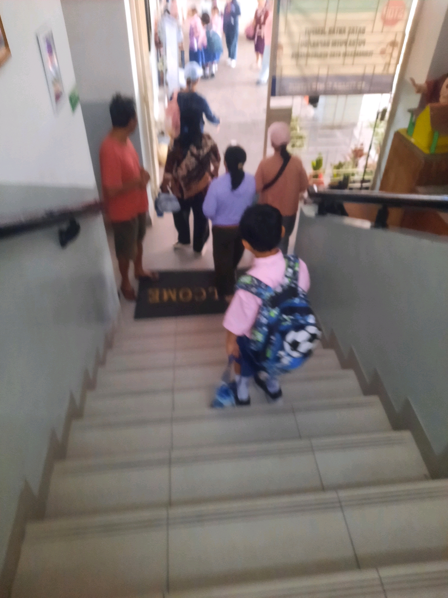 The image depicts a young boy wearing a pink shirt and a blue backpack with a soccer ball design, walking down a tiled staircase. He is holding a blue object in his left hand. Behind him, several adults and children are visible on the stairs and in the background. A woman in an orange shirt and a man in a red shirt are standing near the bottom of the stairs, with a "WELCOME" mat on the floor. Further up the stairs, a person in a purple shirt and a person in a brown and black patterned outfit are present, along with other blurred figures. The environment appears to be an indoor public space, possibly a school or community center, with a banner displayed in the background. The overall scene suggests movement and activity within the location.