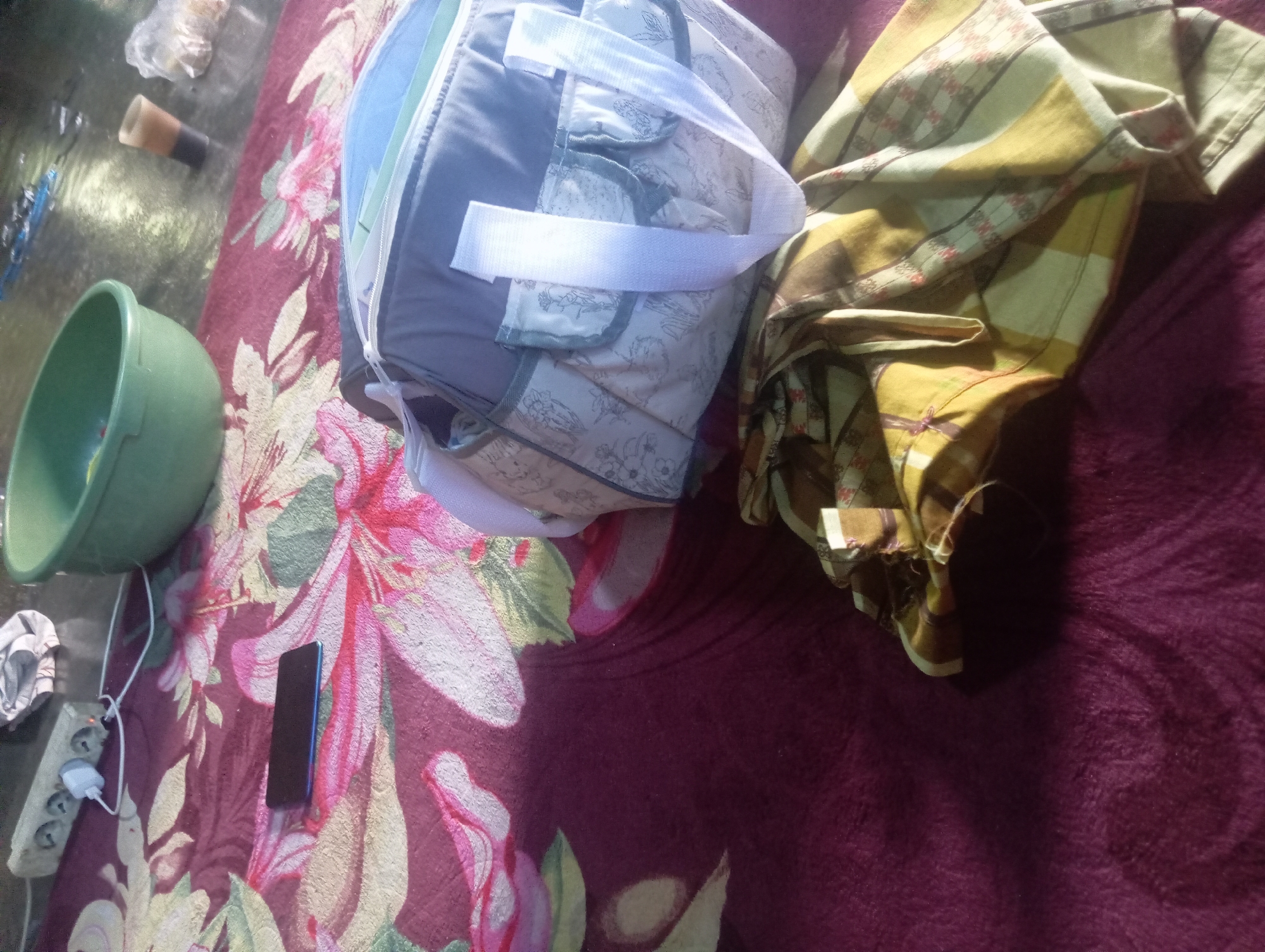 A blanket with a floral pattern in pink, red, yellow, and green is spread on the floor. On the blanket, a black smartphone lies flat. To the right of the phone is a light gray and white diaper bag with a blue lining, featuring a floral pattern in gray and white. Below the diaper bag, a yellow and brown plaid fabric is folded. In the background, a green plastic basin is visible, along with an electrical power strip, a glass with a dark liquid, and a plastic bag. The scene appears to be an indoor setting in Rejosari, Indonesia, possibly a home or a public space where people might rest or store belongings.